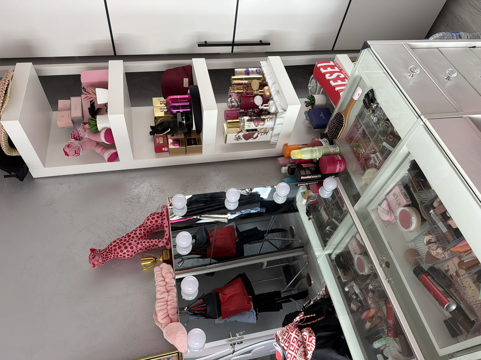 The image depicts a vanity area with a large mirror, shelves, and drawers filled with cosmetics and personal items. The vanity is organized with clear dividers holding makeup, brushes, and accessories. Several perfumes are displayed on the shelves along with decorative items like a pink cheetah statue and a trophy. A makeup artist's chair is reflected in the mirror. The overall setting suggests a personal grooming space, possibly in a bedroom or dressing room. The location is specified as Burgas, Bulgaria.