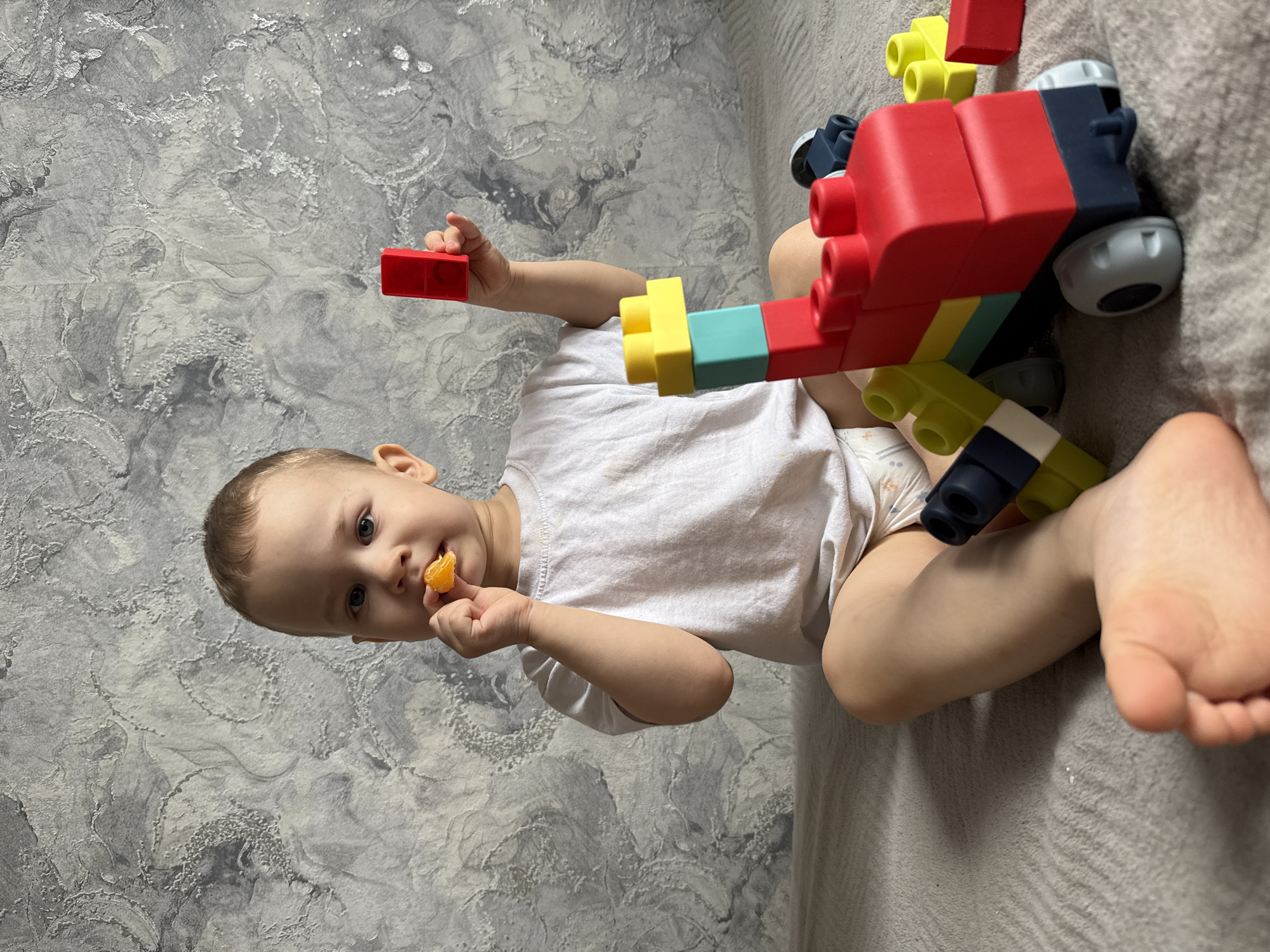 A toddler sits on a light-colored surface, possibly a bed, in a room with a gray textured wall. The child wears a white t-shirt and a diaper. They are eating a segment of an orange with their right hand. The toddler’s left hand holds a red building block. Adjacent to the child, there is a multi-colored building block toy resembling a vehicle. The location is Rusanovo, Russia.