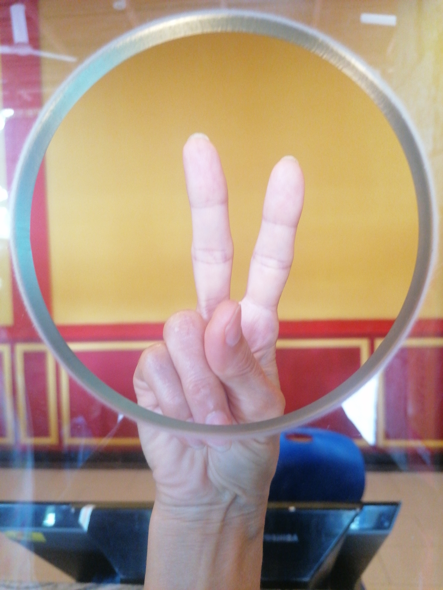 A hand displays a "peace sign" through a round, possibly glass or plastic, barrier. The background appears to be a wall with yellow and red sections, possibly part of an interior architectural design. A dark, rectangular object is visible below the hand, and a blue object is partially obscured behind the barrier. The location is Bai Chay Ward, Vietnam.