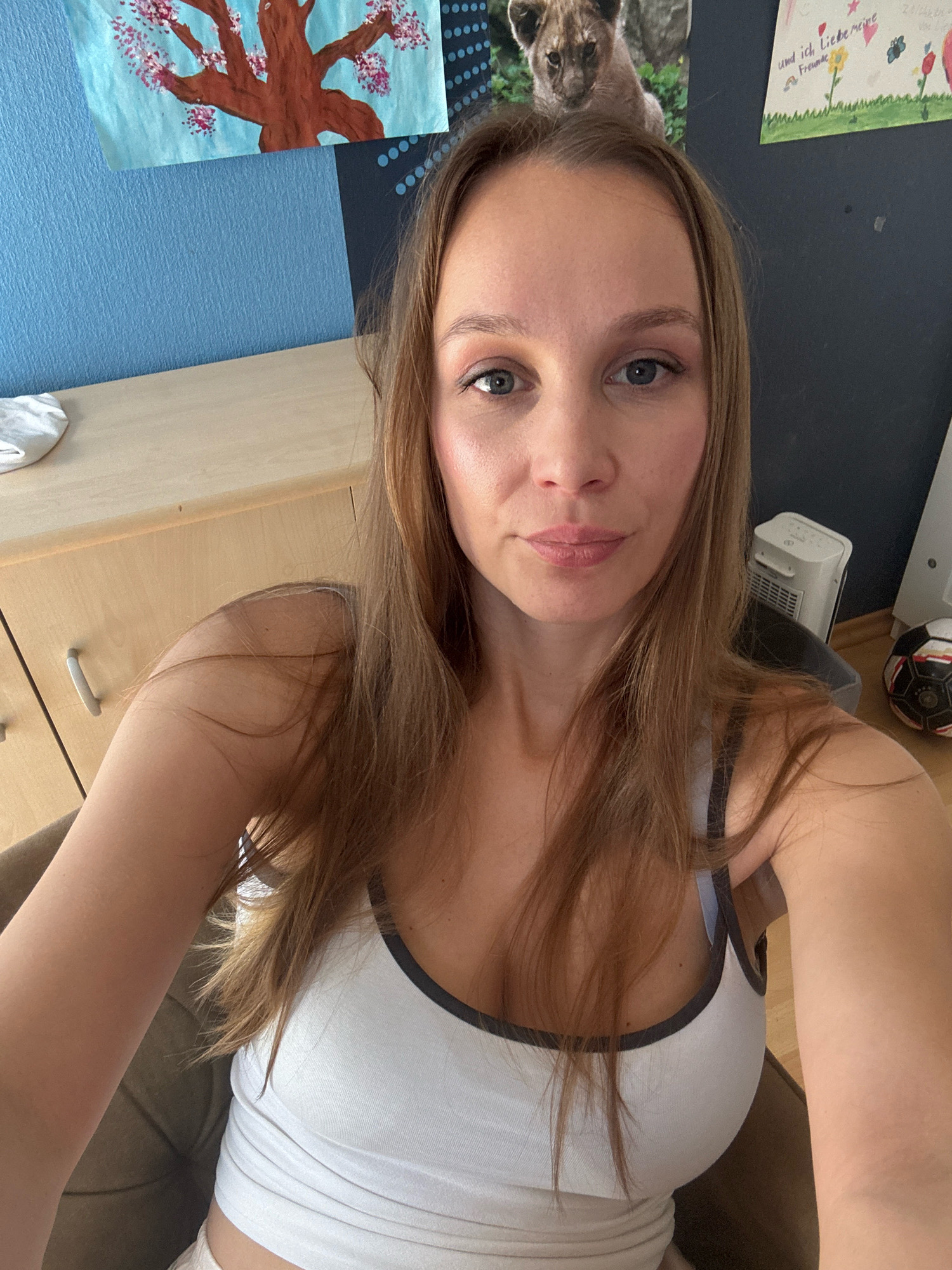 A woman with long, brown hair is taking a selfie in a room. She is wearing a white tank top. In the background, there are two framed pictures on the wall: one of a tree with pink flowers and another with some writing and drawings. There is also a picture of a lion cub on the wall behind her head. To the right, there is a white electric heater and a soccer ball. The woman is sitting in a chair, and the scene appears to be indoors. The location of this room is not specified.