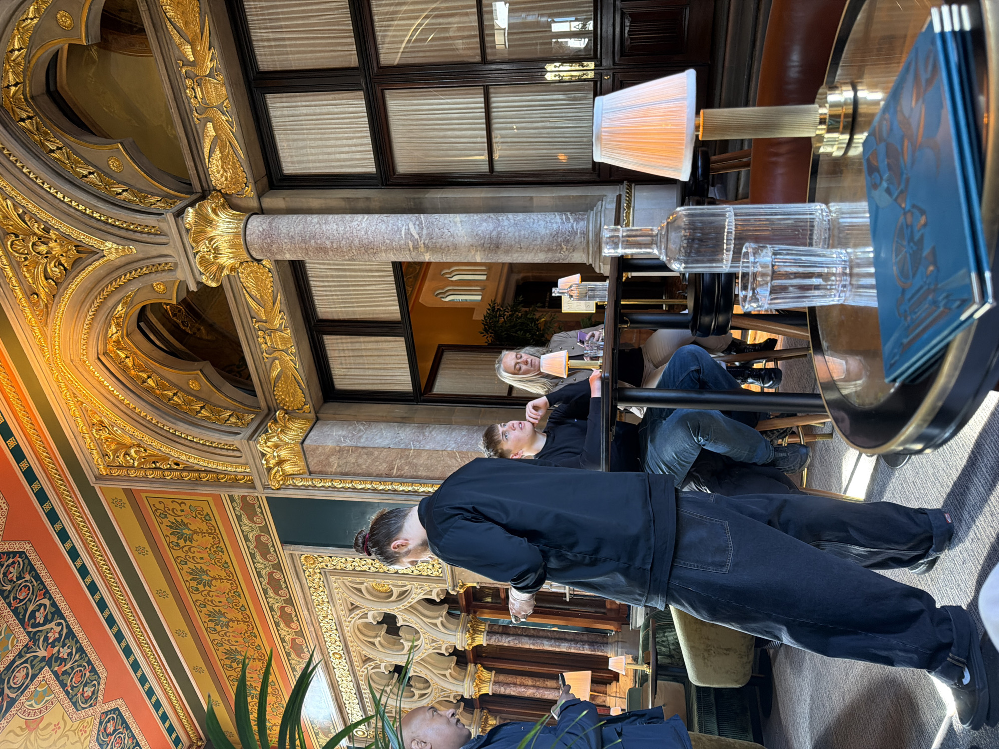 The image captures an opulent interior scene, likely a grand hotel lounge or a historic restaurant, bathed in natural light from large windows in the background. The most striking feature is the highly ornate ceiling and upper walls, richly decorated with gold leaf, intricate carvings, and painted panels in a palette of gold, orange, green, and dark blue. These panels display classical floral and geometric patterns. A prominent marble column with a gilded capital stands on the right side of the frame.
Several individuals are present. In the foreground, a person with dark hair tied in a bun stands with their back to the viewer, wearing a dark jacket and dark jeans. Further back, at a circular table, two people are seated. A blonde woman, partially visible, looks towards the standing person. Opposite her, a man with dark hair is seated, gazing upwards or to the side. The head of another person is visible in the bottom left corner of the frame.
On the table, a clear glass bottle, two empty fluted glasses, and a table lamp with a pleated shade are arranged. A stack of blue menus or booklets, featuring an abstract, possibly geometric pattern, rests on the table. The bright lighting, coupled with the lamps, suggests it is daytime, with the interior ambiance enhanced by artificial light. The general activity appears to be casual social interaction or waiting at a table in a refined setting. No discernible text is visible on any objects. The overall impression is one of elegance and grandeur.