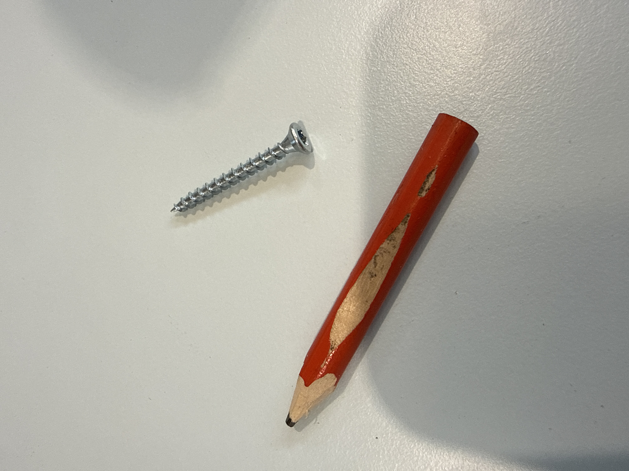 The image shows a red pencil with a sharpened, exposed graphite tip and a metallic screw with visible threading. The pencil is positioned diagonally from the bottom left to the upper right of the frame, with its sharpened end pointing towards the left. The screw is located in the upper right quadrant of the image, also oriented diagonally. Both objects are resting on a light-colored, textured surface, likely a desk or table. There are no people or any indications of a specific location in the city of Fontevivo, Italy. The lighting suggests an indoor environment.