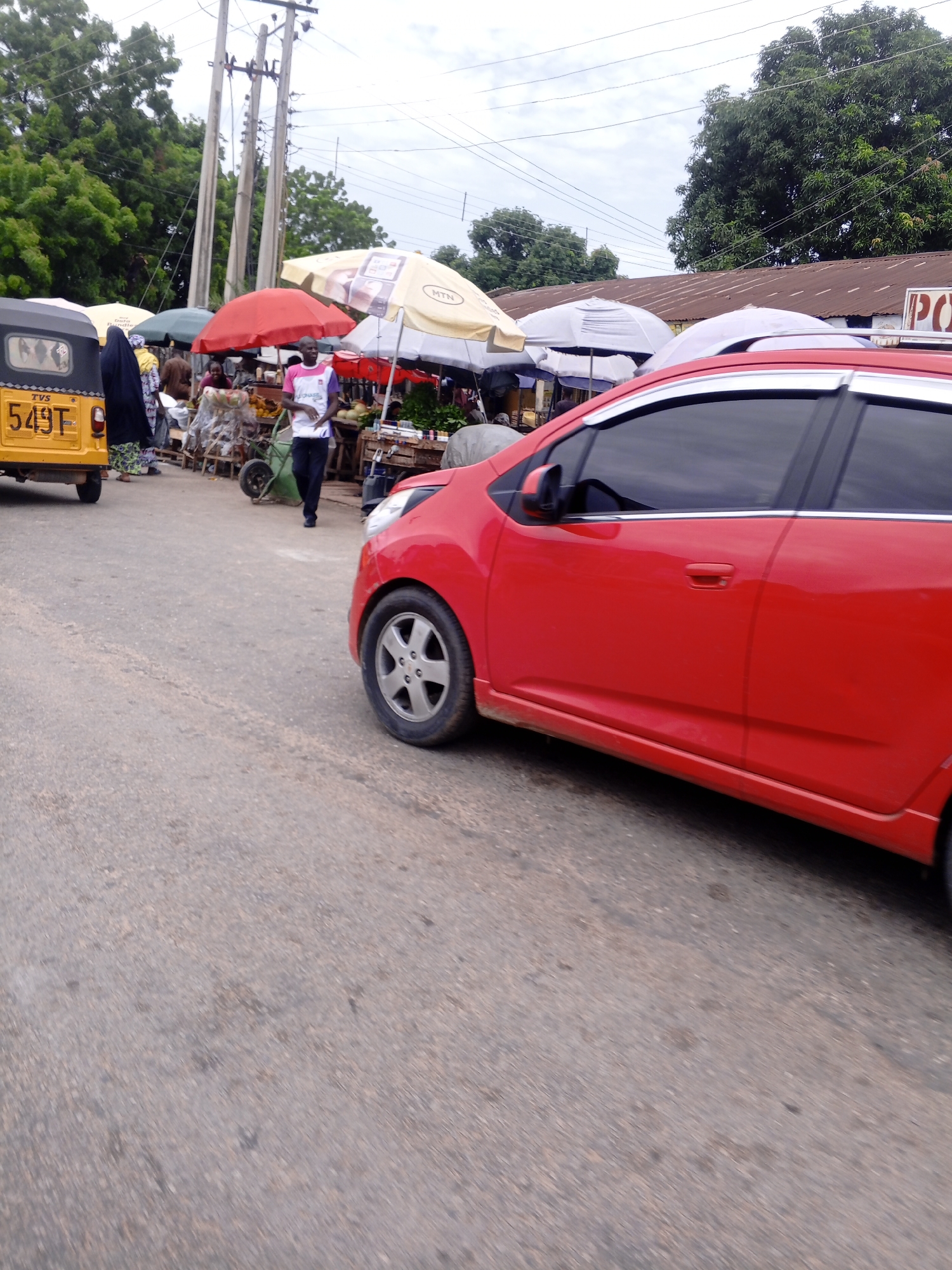 The image captures a street scene in Jalingo, Nigeria, with a focus on a red car in the foreground, moving from left to right. The car's side profile is visible, showing its red paint, tinted windows, and a portion of its front wheel. Beyond the car, a bustling market is depicted. A yellow auto-rickshaw, a common mode of transport, is parked on the left, bearing the license plate "549T". Several people are present, including a person in a dark hijab and patterned skirt near the auto-rickshaw, and another person in a white t-shirt and dark pants walking towards the market stalls. The market features numerous vendors with colorful umbrellas providing shade over their goods, which appear to include fruits and vegetables displayed on wooden crates and carts. In the background, tall utility poles with wires crisscross the sky, and lush green trees line the street. The architecture visible includes corrugated metal roofs. The scene suggests a typical day at a roadside market in Jalingo.