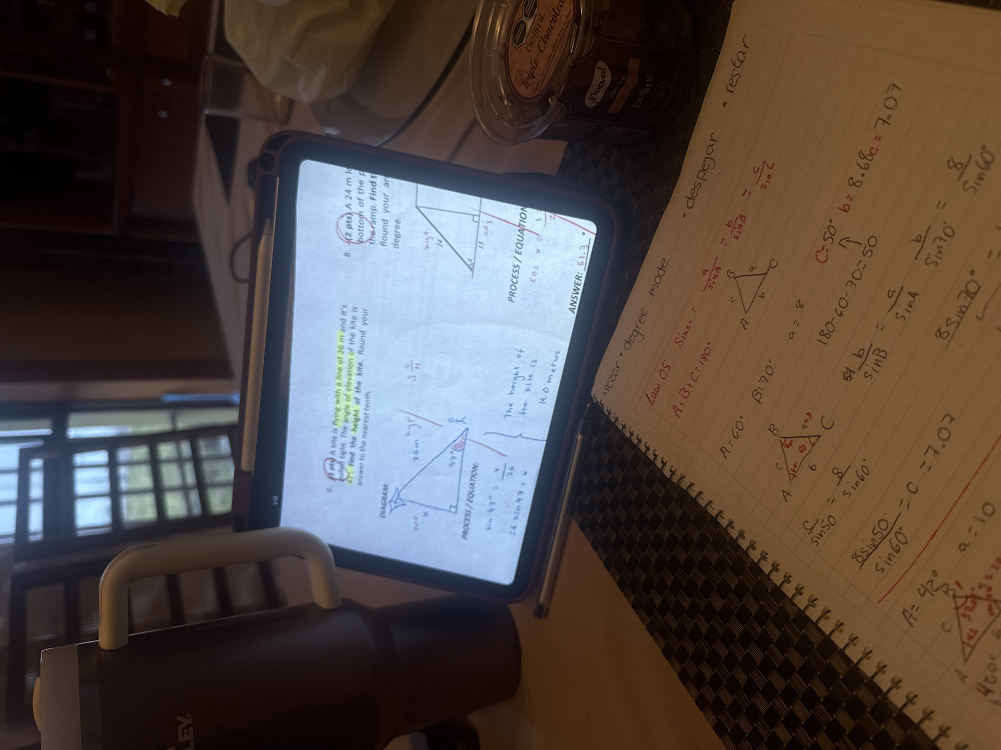 A slightly high-angle, medium shot shows a dark-colored desk or table mat upon which a tablet, a notebook, a jar of Bellini Triple Chocolate dessert, and a dark-colored Stanley brand beverage container are positioned. The tablet, propped up in a case, displays what appears to be math problems related to trigonometry involving kites and ramps. A stylus is seen next to the tablet. Adjacent to the tablet, a spiral-bound notebook is open, displaying handwritten math equations and diagrams, again seemingly related to trigonometry, including the Law of Sines and triangle calculations. The pages include annotations in what appears to be Spanish. The container of Bellini Triple Chocolate is partially visible in the upper right corner. In the bottom left corner, a portion of a dark-colored Stanley beverage container is visible. The background is blurred, but a window or doorway is visible, suggesting an indoor location.