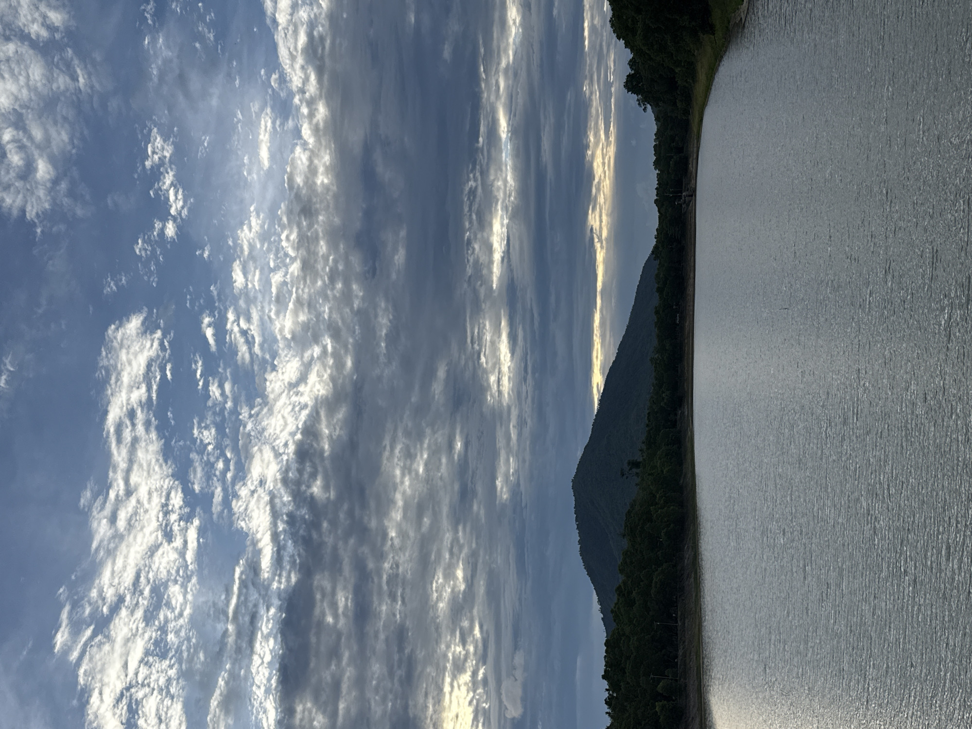 The image depicts a serene natural landscape, likely a reservoir or a large body of water. In the foreground, the water surface is calm with slight ripples, reflecting the muted tones of the sky. A dense line of dark green trees borders the water on the left and right sides. Behind the trees, a prominent, rounded mountain covered in vegetation rises against the sky. The sky itself is dramatic, filled with a multitude of clouds. Some clouds are white and wispy, catching the light, while others are darker and more textured, suggesting an overcast or approaching storm. The overall lighting suggests either early morning or late evening. The location is identified as Ko Samui District, Thailand, suggesting a tropical or subtropical environment. There are no people or human activity visible in the image, emphasizing the natural and tranquil atmosphere.