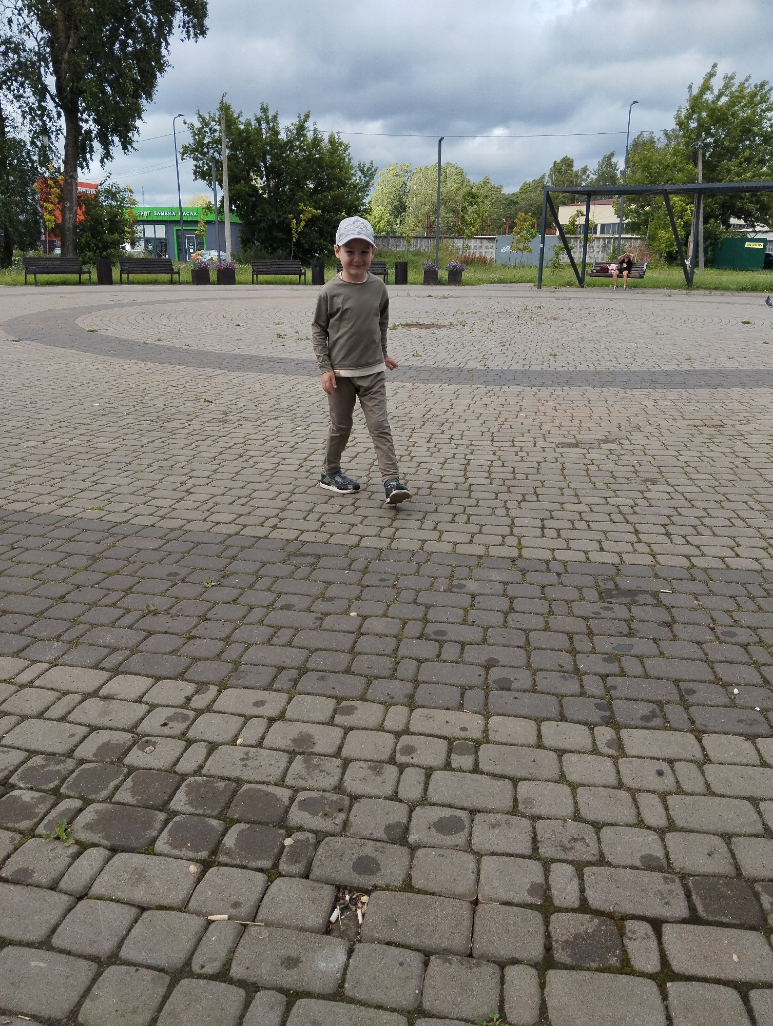 A young boy wearing a light gray long-sleeved shirt, khaki pants, and a light-colored baseball cap stands on a paved courtyard, looking towards the camera. He has a neutral expression. The courtyard is paved with grey cobblestones and has circular patterns. In the background, there is a park-like setting with trees, benches, a building with green and gray siding, and a swing set. The sky is overcast with dark clouds. The time of day appears to be afternoon. The boy is positioned in the foreground, slightly to the right of the center, and is in focus. The background elements, including the building and swing set, are slightly out of focus. There is some litter on the ground in the lower right of the image, including cigarette butts. There is text visible on the building in the background, which reads "SPORT ЗАМЕНА АСАА".