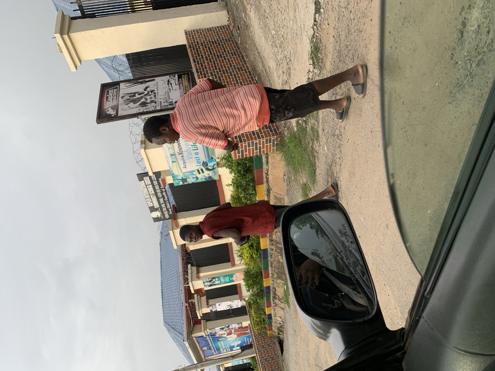 The image is taken from inside a car with the side mirror visible. The scene appears to be on a street in Abuja, Nigeria. A man in a red and white striped shirt and dark shorts is standing on the side of the road. The side of a building is visible with multiple signs displayed on the front. One sign displays "KAPLEN INTERNATIONAL COMMUNITY SCHOOL (KICS)" and lists phone numbers. Another sign references a business center and the phrase "Acquire Knowledge Values Last a Life."