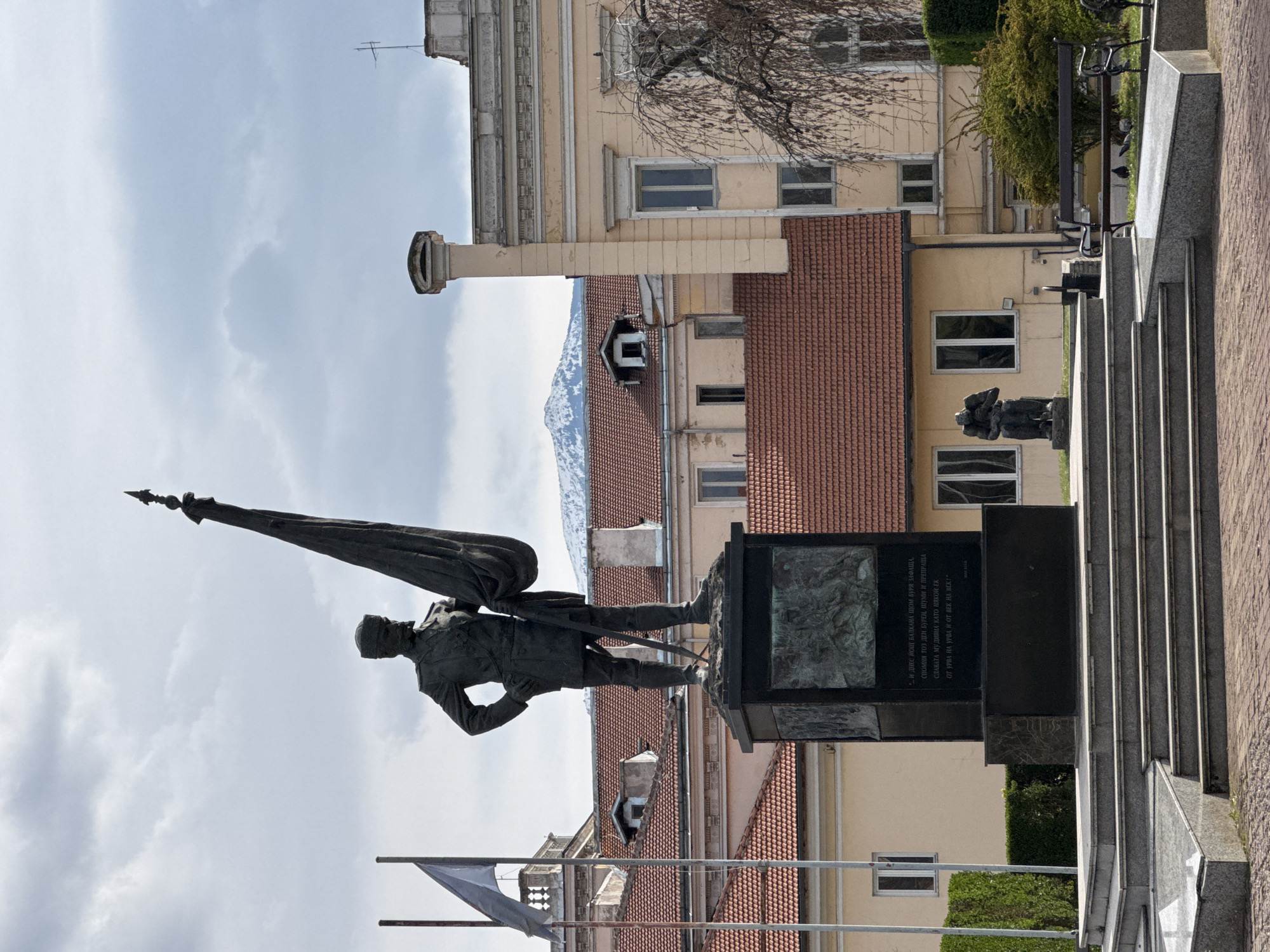The image shows a bronze statue of a soldier carrying a flag, standing on a stone base with inscriptions in Bulgarian Cyrillic. In the background are buildings with beige and brown tile roofs, and beyond that, a mountain range. The statue and buildings appear to be located in an urban setting, specifically the city of Sofia, Bulgaria. The sky is overcast. In addition to the buildings, there is also a glimpse of park benches with shrubbery behind them.