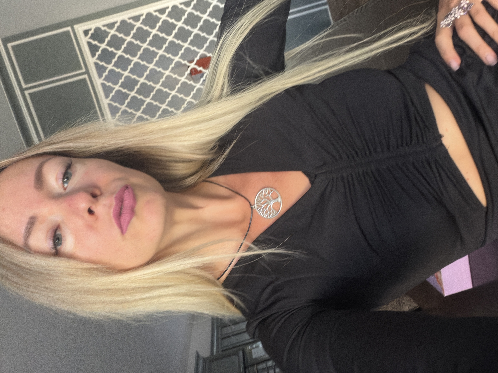 A woman with blonde hair, wearing a black top and a silver tree pendant, takes a selfie indoors. The background features a grey wall with decorative white patterns and what appear to be kitchen cabinets. Her makeup includes pink lipstick and eyeliner. She is wearing a ring on her right hand. The image does not contain any information that would place the scene in Xuân Trường - Đà Lạt, Vietnam.