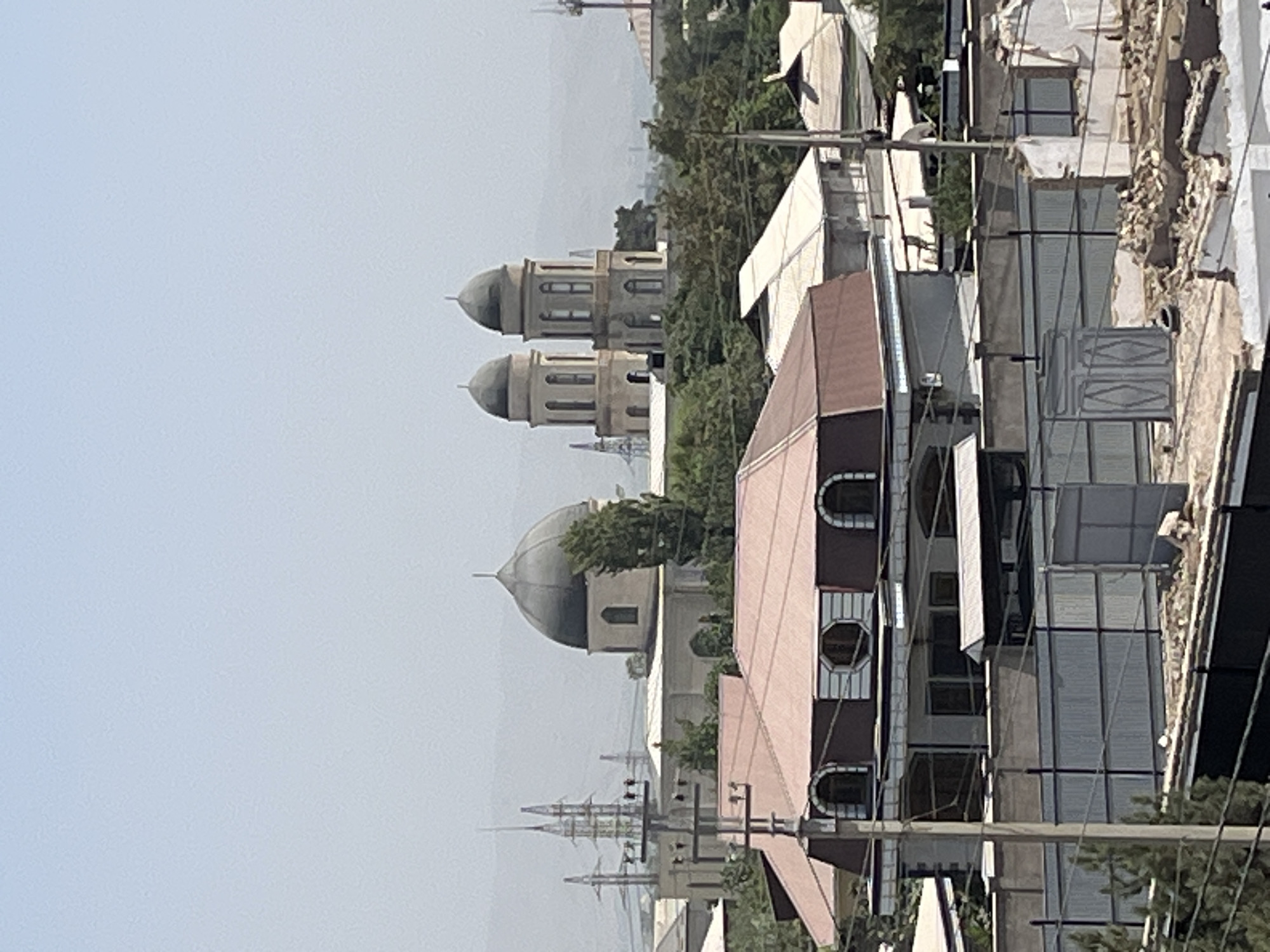 This image shows a cityscape with a prominent building featuring a large dome and two smaller domes, possibly a mosque or religious structure, in Bokhtar, Tajikistan. In the foreground, there are several residential buildings with pitched roofs. Power lines and transmission towers are visible in the background, indicating urban infrastructure. The sky is hazy and light blue, suggesting a daytime scene, possibly with dust or pollution in the air. There are no people clearly visible in the image. The overall scene depicts a mix of religious and residential architecture within an urban environment.