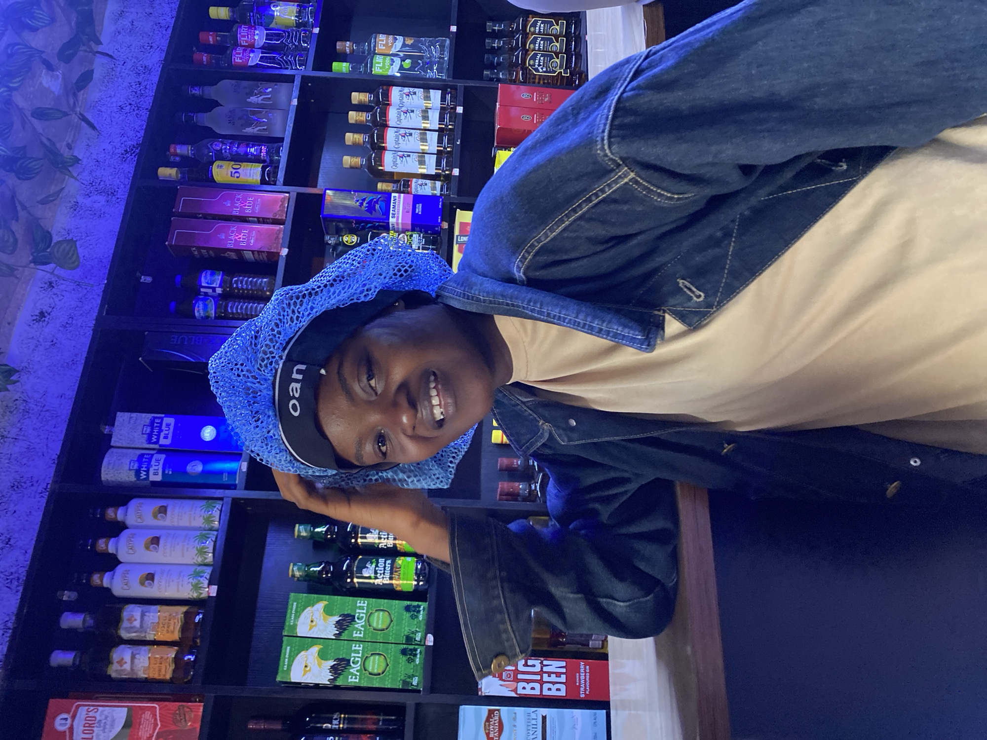 A young woman wearing a blue hair net and a denim jacket is standing behind a bar counter in a dimly lit establishment. The bar counter is made of wood. Behind her, shelves are stocked with various bottles of alcoholic beverages. The bottles appear to be of different brands and types, including spirits, liqueurs, and potentially wines, with labels such as "Lord's," "Calypso," "White Blue," "Eagle Eagle," "Action Bitters," "Black Label," "Captain," and "Flirt Vodka." The lighting in the establishment is predominantly blue and appears to come from overhead fixtures and possibly the illuminated shelves. The background wall has a textured, possibly leaf-like pattern, and is illuminated by the blue light, creating an atmospheric effect. The woman is looking towards the camera and smiling. The overall scene suggests a retail liquor store or a bar. The specific location in the city of Bar Jirgi Summa, Nigeria, cannot be determined from the visual information provided in the image.