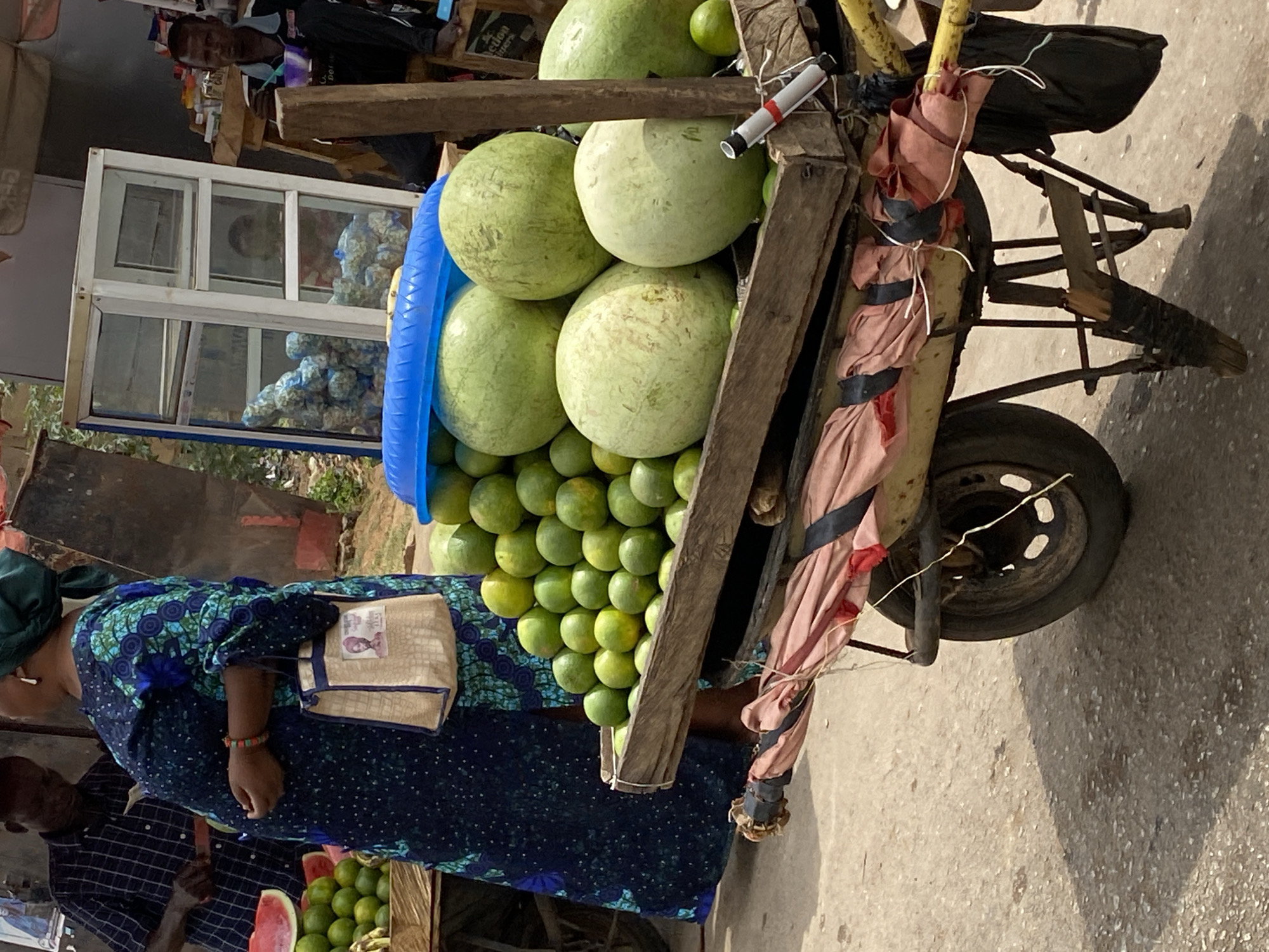 A mobile fruit vendor operates in Bukuru, Nigeria. The vendor uses a cart with a single wheel and wooden supports. Stacked on the cart are several large, light green fruits, possibly gourds, above a layer of smaller green fruits, likely limes. To the left of the cart stands a woman in a blue patterned dress, a beige purse at her hip. Beside her is a man in a plaid shirt. In the background is a glass display case, with other individuals visible nearby.