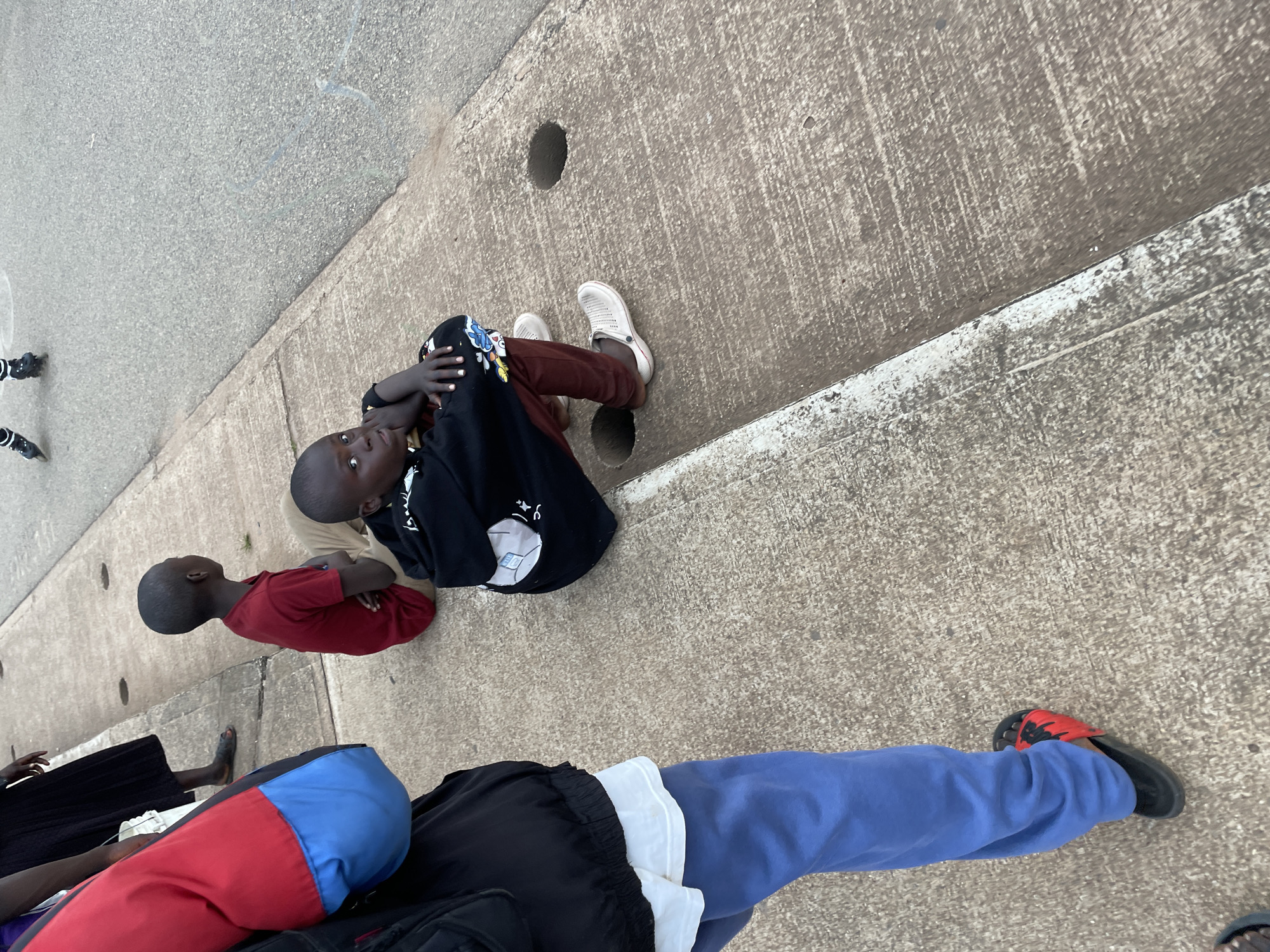 The image shows a group of children sitting on a sidewalk in Jos, Nigeria. The children appear to be waiting for something. In the foreground, a young boy is sitting facing the camera, with his arms crossed. He is wearing a black hoodie and brown pants. To his left, another boy is sitting with his back to the camera, wearing a red t-shirt. In the background, a woman is standing, wearing a black dress. The sidewalk is made of concrete, and there is a road to the right of the sidewalk. There are also some drain covers on the sidewalk. The overall scene is one of everyday life in a Nigerian city.