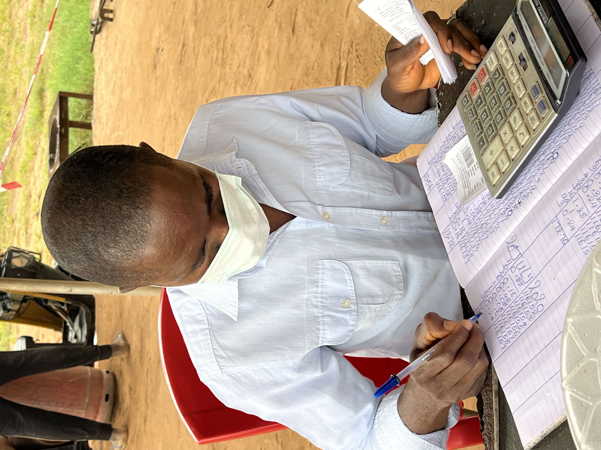 A Black male wearing a light blue pinstripe button-down shirt and a light blue face mask is seated at a red chair outdoors, focused on a lined notebook and a calculator. He is writing in the notebook with a blue pen in his right hand, while holding a slip of paper in his left. The notebook is open to a page filled with handwritten numbers and calculations. A calculator with a digital display sits on the notebook. The background shows a sandy area with a wooden table, some grass, and caution tape. In the far background, parts of a person in dark pants and shoes, and a yellow vehicle are visible. The scene appears to be in Nkpor, Nigeria.