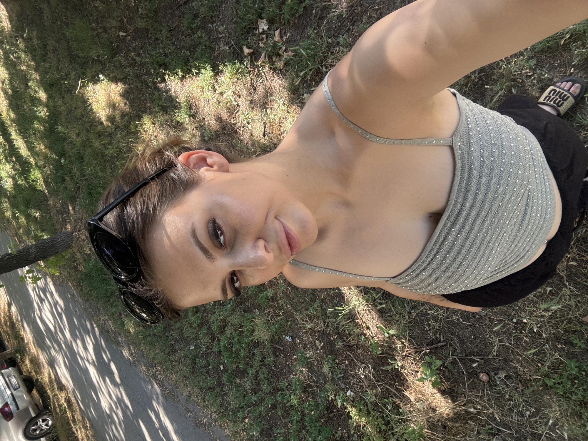 The image shows a woman taking a selfie outdoors. She is looking towards the camera with a slight smile. She has dark hair pulled back and is wearing black sunglasses perched on her head. Her top is light gray and appears to be embellished with small, reflective dots. She is also wearing black shorts. Her left foot is visible, wearing a black slide sandal with a tan sole and the "MIU MIU" logo.
The setting is a grassy area next to a paved path. Sunlight filters through trees, casting dappled shadows on the ground and path. A portion of a light-colored car is visible on the left side of the path, with a tree trunk on the far left. The ground is covered with dry grass, dirt, and some scattered leaves and twigs, suggesting it might be a park or a natural area.
The lighting indicates it is daytime, likely late morning or afternoon, given the angle of the shadows. The weather appears to be warm and sunny. The overall mood is relaxed and casual. No discernible text is visible other than the brand logo on the sandal. The location context of Plovdiv, Bulgaria, suggests an outdoor urban or semi-urban environment.