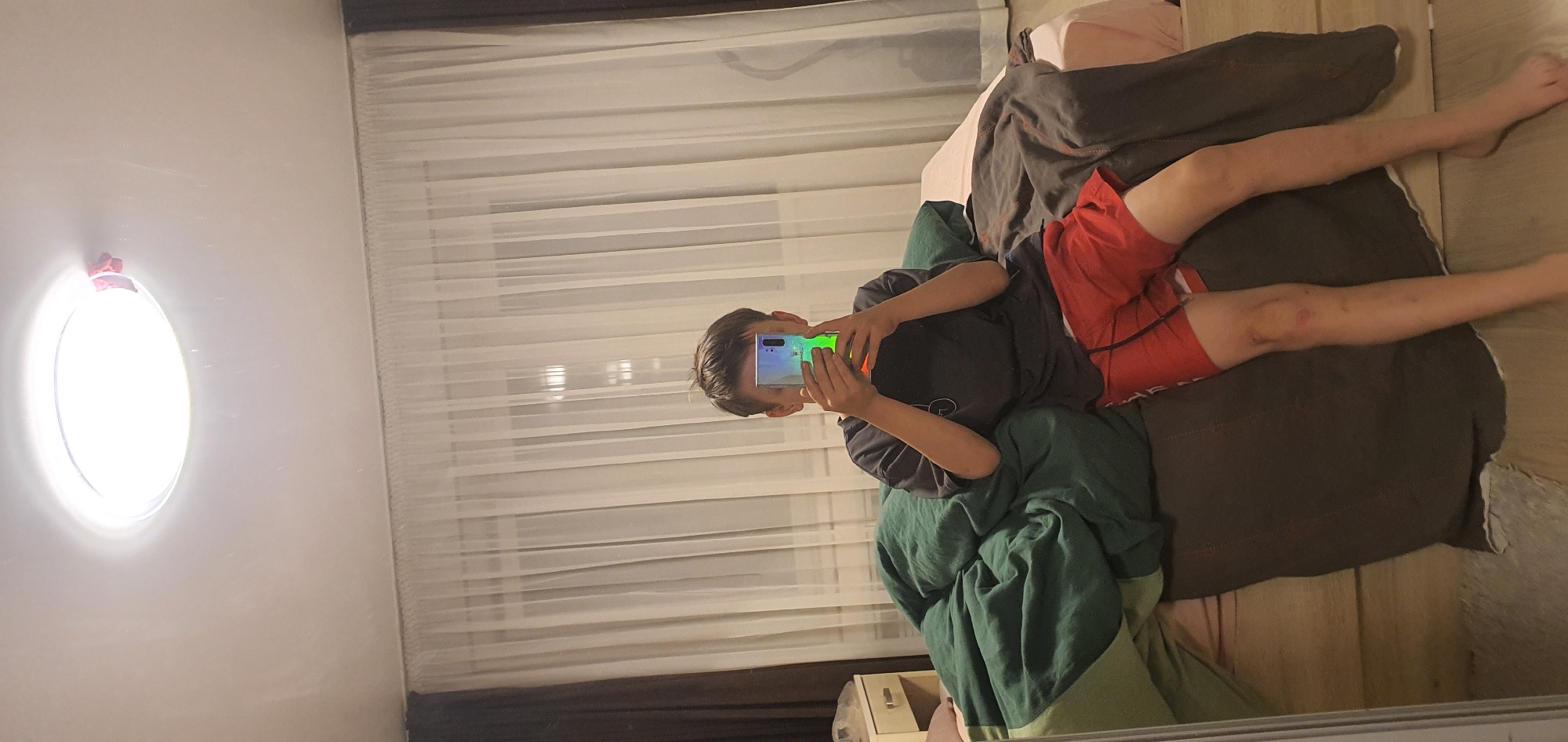A young person is lying on a bed in a room with light-colored walls, a white curtain, and a round window. The person is holding a smartphone with a bright screen, obscuring part of their face. They are wearing a dark t-shirt and red shorts. Bedding includes green, brown, and pink covers. The exact location within Chaikovsky, Russia, cannot be determined from the image.