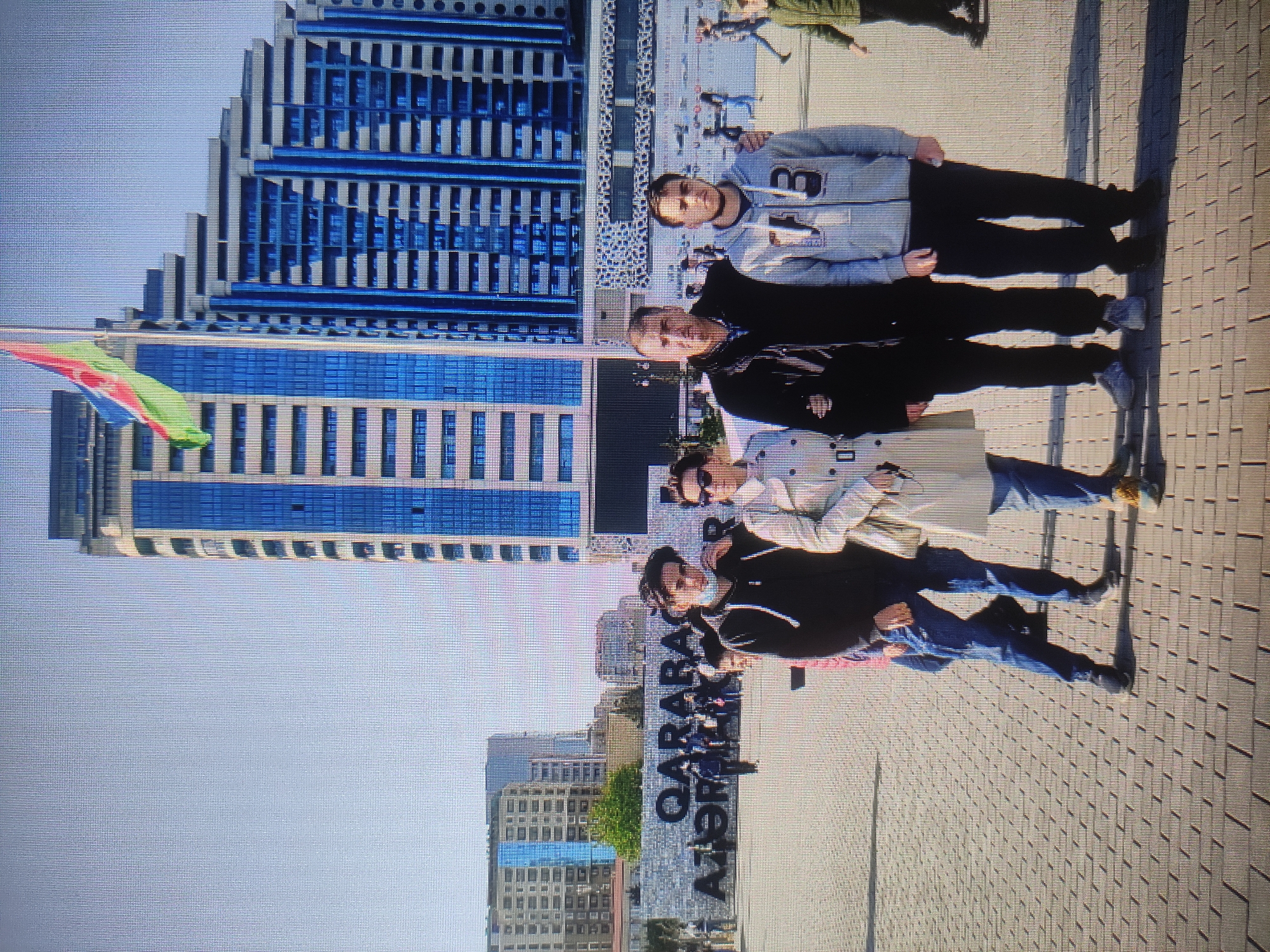 The image depicts a family standing on a paved plaza in front of a large, modern building with blue-tinted windows. A flag, predominantly green, red, and blue, is visible at the top of the building. In the background, large letters spelling out "QARABAĞ AZƏRBAYCAN" are present, indicating the location as Baku, Azerbaijan. The family consists of two adults and two young men. The adult man on the left has his arm around the shoulders of the young man next to him. The adult woman is on the far left. The young man on the far right is wearing a grey hoodie with the number "73" on it. The weather appears to be sunny.