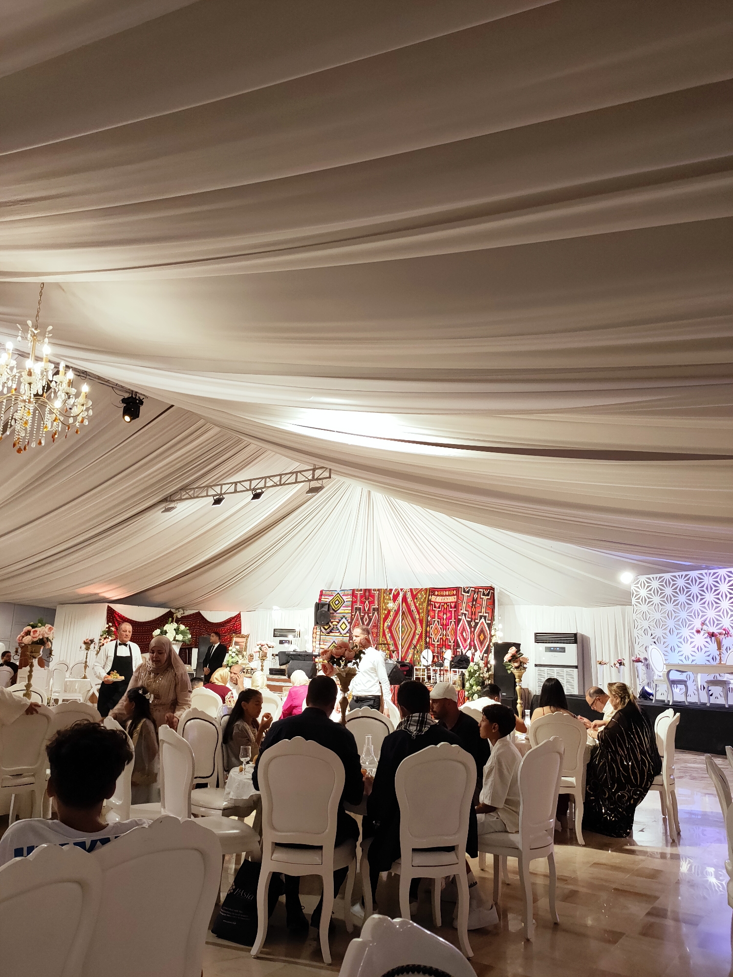 This image depicts an indoor event, likely a wedding reception or similar celebration, taking place within a large tent structure in Mornag, Tunisia. The ceiling is draped with white fabric, creating a formal and elegant atmosphere, with a crystal chandelier visible on the left. Guests are seated at round tables, predominantly dressed in formal attire.
In the foreground, several guests are seated, with a young person in a white shirt facing away from the camera. To the right, a woman in a black sequined dress is engaged in conversation with a man. The middle ground shows more guests seated at tables, with a notable group including children. A man in a white shirt and black apron, possibly a server, is interacting with a woman wearing a hijab. Behind them, a large tapestry with geometric patterns hangs on the wall.
The background features additional seating, floral arrangements, and decor, including a white wall with a geometric pattern on the far right. An air conditioning unit is visible on the back wall. The overall impression is of a well-attended, celebratory gathering in a decorated indoor venue.