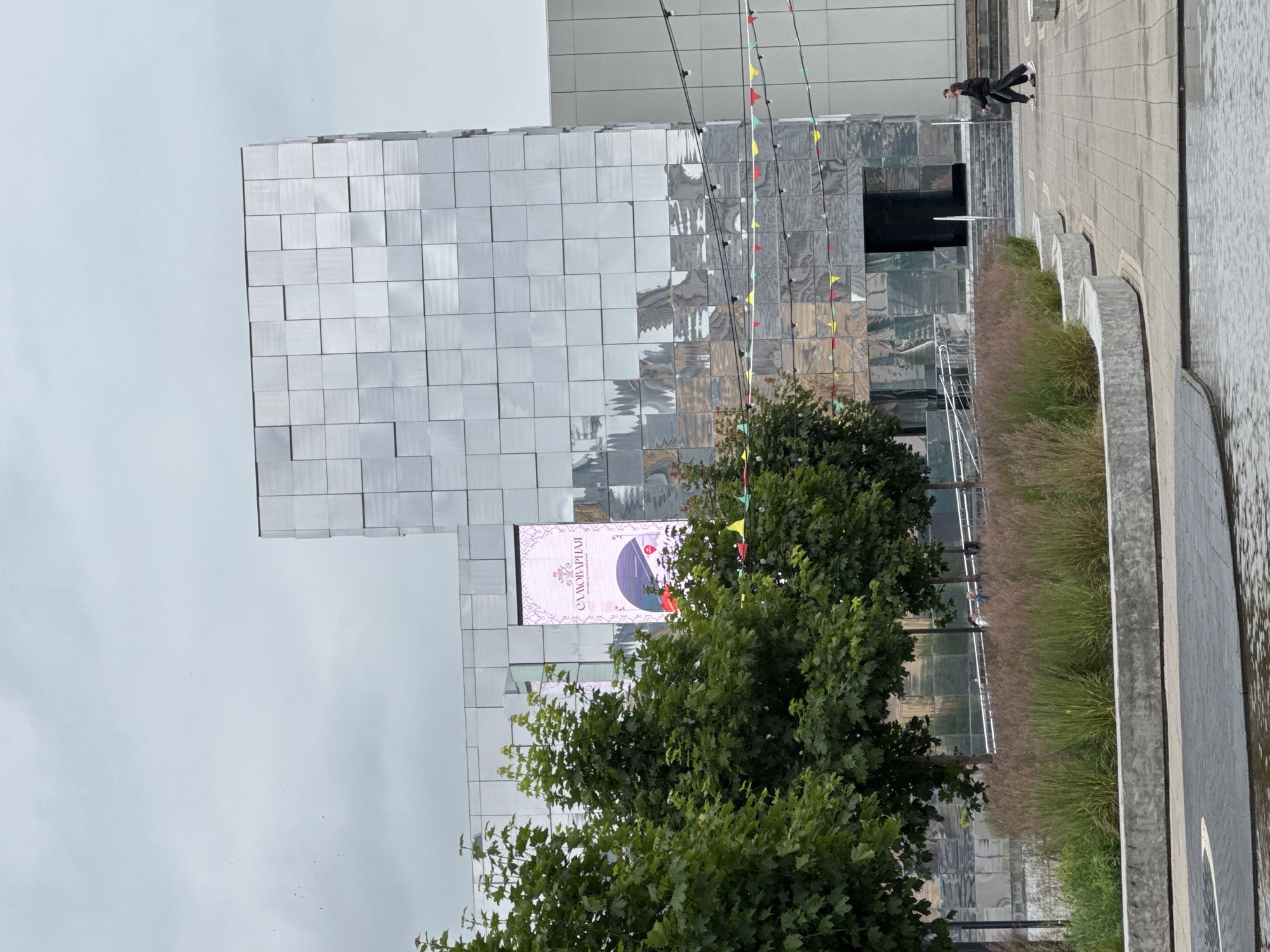 The image shows the facade of a modern building in Nizhny Novgorod, Russia. The building is clad in reflective, square panels, giving it a striking, geometric appearance. A large digital screen on the building displays an advertisement for "Самоварная" (Samovarnaya), described as a restaurant of Russian cuisine. In front of the building, there are several trees with green foliage and a landscaping area with dry grasses and low concrete planters. A paved walkway is visible in the foreground, leading towards a reflective pool of water. Two people, a man and a woman dressed in dark casual clothing, are walking along the paved area away from the building and towards the right side of the frame. The sky is overcast and grey.