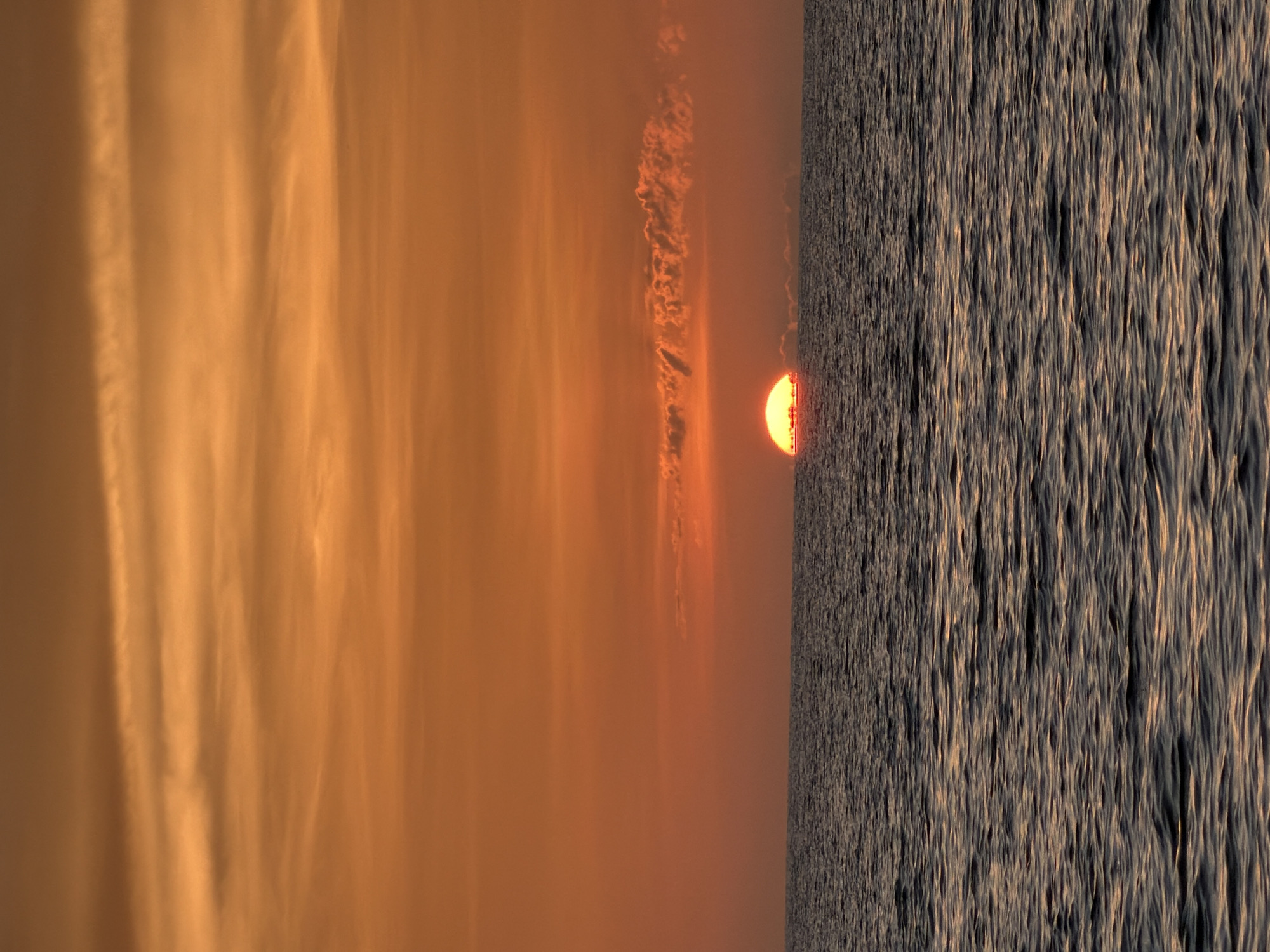 An extreme wide shot captures a sunset over the ocean in Ko Samui District, Thailand. The sun, a bright orange disc, is half-submerged on the horizon, casting a warm glow across the sky. The sky is filled with wispy, orange clouds that stretch horizontally across the frame. The ocean below is a deep blue with gentle waves, reflecting the colors of the sunset. The overall scene is serene and tranquil, evoking a sense of peace and natural beauty.