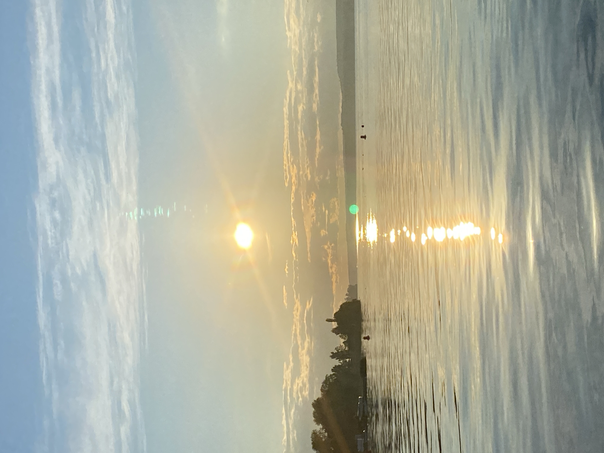 The image captures a serene lake scene at sunrise, presumed to be in Votkinsk, Russia. The sun, a bright orb with visible lens flare, hangs in the hazy sky, casting a golden reflection across the rippling water. Wispy clouds are scattered across the blue expanse. On the left, a densely wooded shoreline is visible, with a few boats docked and a red buoy in the water. The distant horizon is marked by a low, tree-covered ridge. The overall atmosphere is tranquil and calm, with the gentle movement of the water and the soft light of dawn.