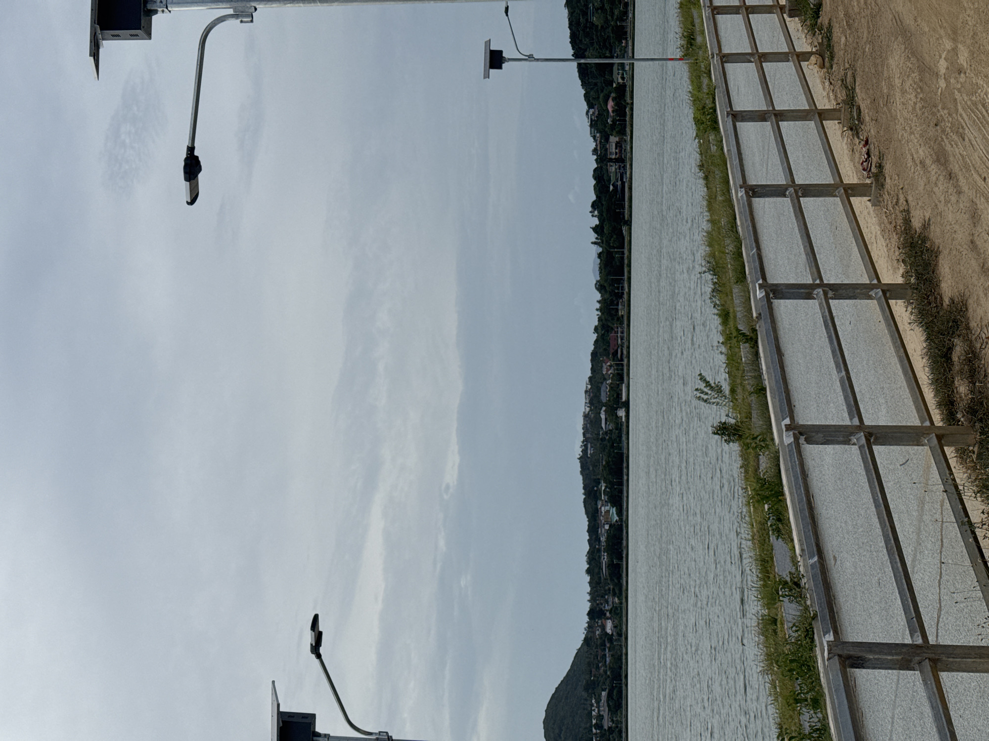 This is an outdoor, medium-shot, vertical photograph taken on a cloudy day. The scene depicts a scenic view of a body of water, likely a bay or a large lake, with a distant shoreline populated by trees and buildings. A paved walkway with a metal railing runs along the foreground, parallel to the water. Several solar-powered streetlights are visible, two of them prominently in the frame. The water exhibits a textured surface with small ripples. The distant land features a hilly landscape with vegetation and scattered houses. The sky is overcast with diffused light, suggesting a possibly humid atmosphere common in the region of Ko Samui District, Thailand. There are no people or animals visible in the image.