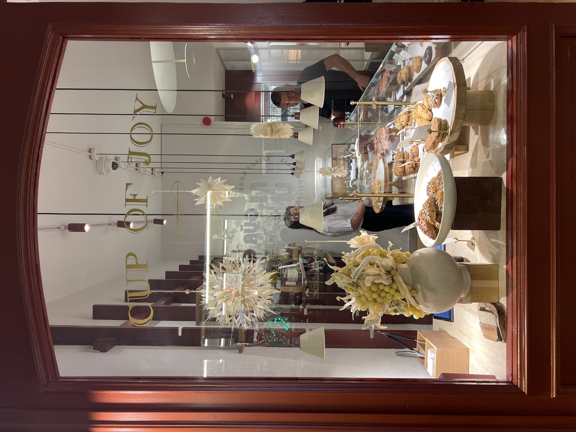 A shop window displays "CUP OF JOY" in gold lettering. Inside, a woman in a white shirt and a man in a black t-shirt are working behind a display case filled with pastries and baked goods. The man is holding a dark object and appears to be attending to the display. The shop window has a dark red frame. A decorative arrangement of dried yellow foliage is in a light-colored vase in the foreground, positioned on a wooden block. Behind the vase, a small wooden box holds tongs and napkins. The shop appears to be a bakery or cafe located in Beşiktaş, Turkey.