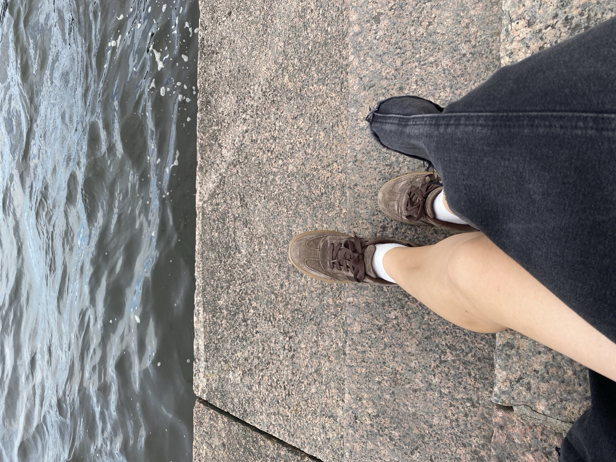 The image shows a person's legs and feet resting on a granite embankment overlooking a body of water. The person is wearing brown sneakers and white socks, and dark-wash denim jeans. The water appears calm with small ripples on the surface. The granite embankment is made of rough, speckled stone. The scene is likely located along the Neva River or one of its canals in Saint Petersburg, Russia, given the characteristic granite embankments found throughout the city.