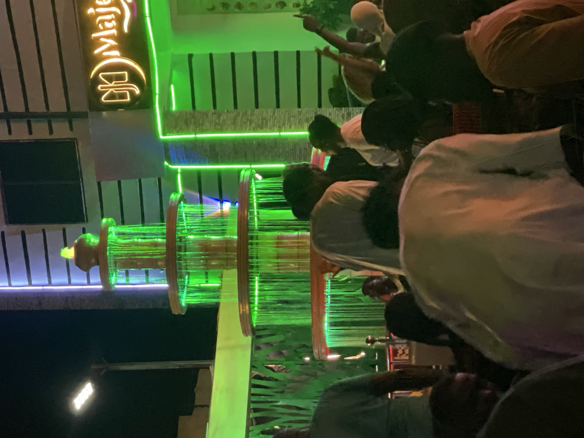 The image shows an outdoor scene at night. A building with a neon sign "Majesty" is visible on the left side. A tiered fountain-like structure is illuminated with green lights. Several people are present, some with their backs to the camera, creating a sense of a crowded or bustling environment. The overall setting appears to be in Tumfure, Nigeria, based on the context. Details about individual actions or interactions between people are limited due to the perspective and focus on the illuminated structures.