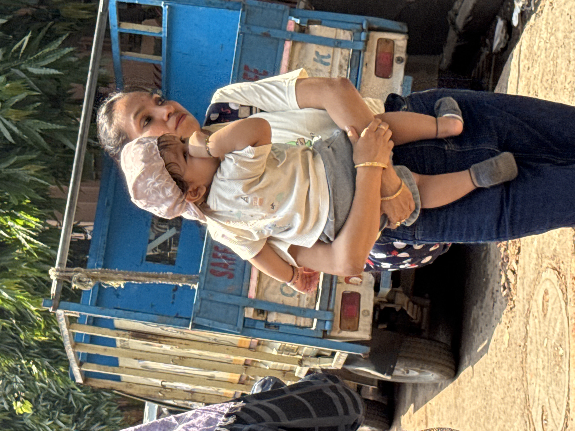 The image shows a woman in blue jeans holding a baby dressed in light colors in front of a blue truck. The location is inferred to be Kolkata, India, based on the style of the truck and the appearance of the individuals. The woman is wearing gold bangles on her wrists. The baby has a head covering. They appear to be on a street. A tire of the truck is visible in the background, and the surrounding environment suggests an urban setting.