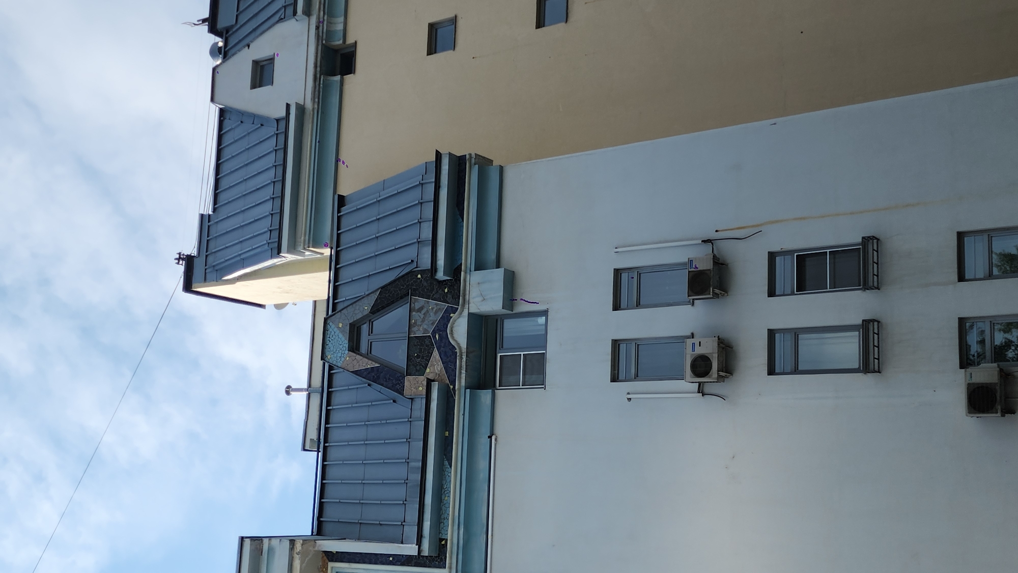 The image shows a close-up, low-angle view of the upper floors of two adjacent buildings. The building in the foreground is lighter in color, possibly white or light gray, with a light blue trim along the roofline. It has several windows and air conditioning units visible. The building in the background is a light yellow or beige color and appears to be taller. Both buildings have dark gray metal roofs with visible seams. One roof has a distinctive triangular section with mosaic tile detailing. The sky above is blue with scattered white clouds. The scene appears to be an urban environment, likely in Moscow, Russia, given the architectural style and common use of air conditioning units on buildings. There are no people visible in the image, and no discernible actions or interactions taking place.