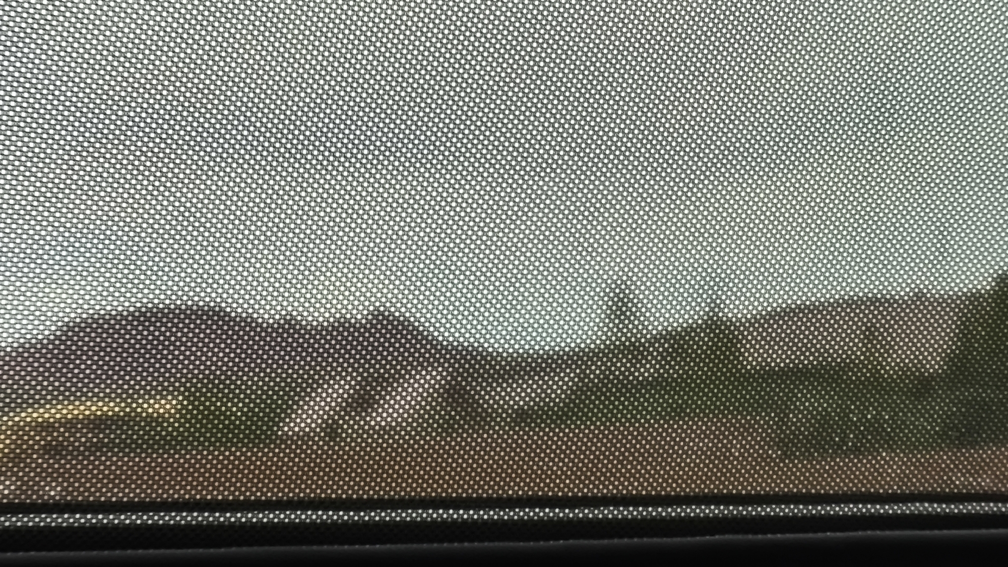 The image, taken on May 30, 2024, at 5:55 PM MDT, shows a view through a mesh screen, likely a window screen or sunshade, of an outdoor scene. The foreground is dominated by the circular pattern of the mesh. Beyond the screen, a landscape is visible, featuring a blurry row of buildings or houses, interspersed with trees and vegetation. The overall lighting suggests an overcast or hazy day, with muted colors in the background. The location is specified as Virgin, Utah, United States of America, although no specific landmarks are identifiable within the image to pinpoint a precise spot within the city. There are no people or animals visible. The scene appears to be a residential or semi-rural environment.