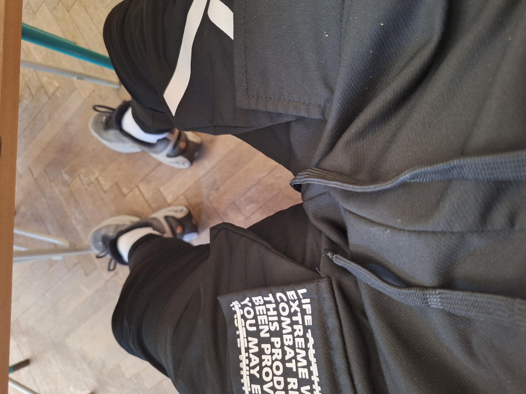 The image shows a first-person perspective of a person wearing black sweatpants with white text and a white stripe on the left leg, and athletic shoes. The person is sitting at a wooden table, with their legs crossed and resting on a wooden floor with a parquet pattern. In the background, there are metal chair legs. The scene appears to be indoors, possibly in a classroom or a public space with tables and chairs. There is no visible indication of the location being in the city of Varna, Bulgaria.