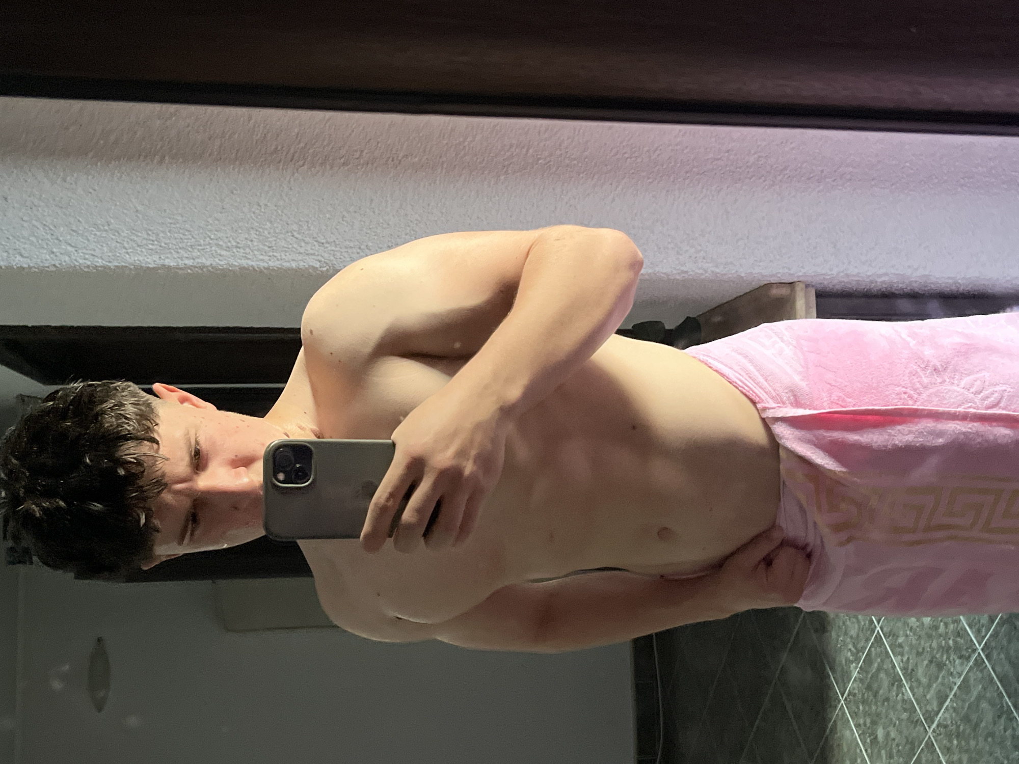A shirtless young man stands in a bathroom with light gray walls and dark tile flooring, possibly taking a selfie using a smartphone. He is partially covered by a pink towel with a geometric pattern. The location is assumed to be in Zlatograd, Bulgaria.