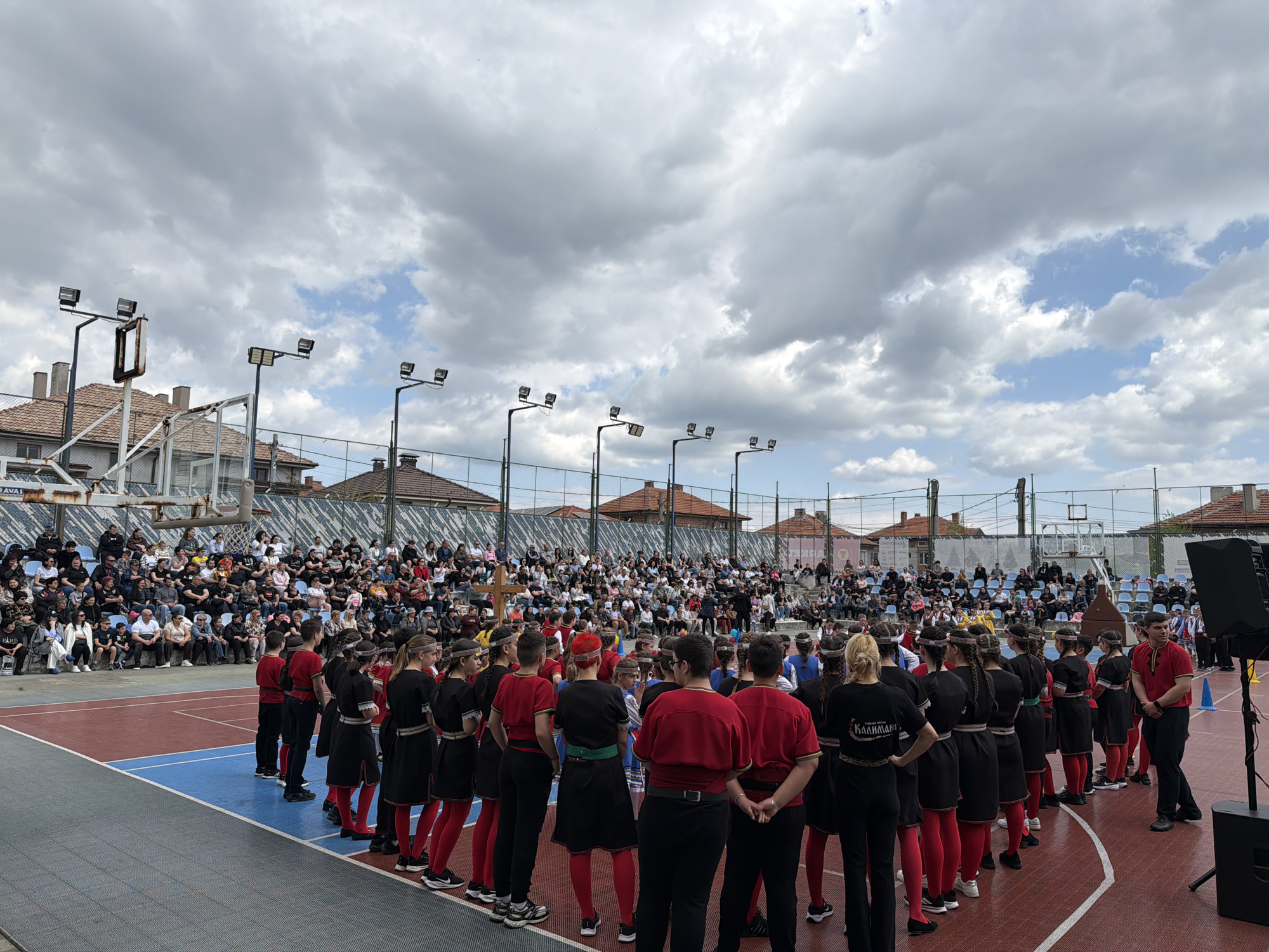 A large group of people is gathered on an outdoor basketball court, likely in the city of Rakovski, Bulgaria. In the foreground, a line of people wearing traditional clothing (red shirts, black tunics, and red leggings) stand with their backs to the camera, facing a crowd seated in bleachers beyond the court. The crowd appears to be watching the group in traditional clothing. Above the court, overhead lights are visible, along with buildings featuring red tile roofs. The sky is overcast with clouds. A basketball hoop is visible on the left side of the image. A black speaker is present on the right side of the image.
