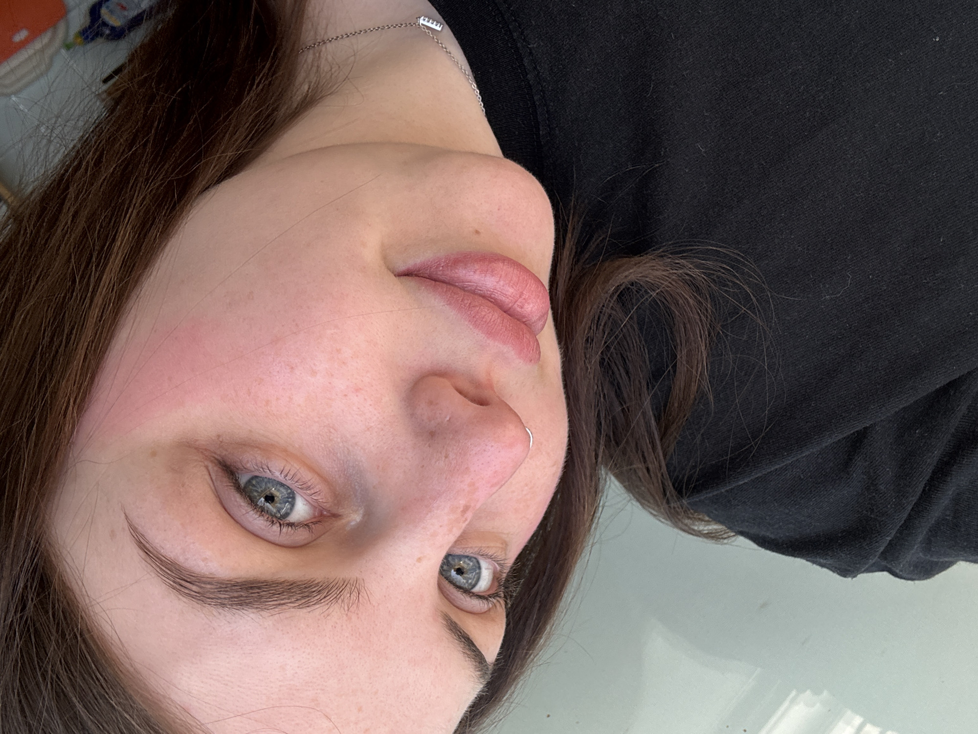A close-up, upside-down portrait shows a Caucasian woman with fair skin, light blue eyes, and brown hair. She has a silver nose ring in her left nostril. The woman is wearing a black top and a delicate silver necklace. Her lips appear to be slightly parted. Freckles are visible on her face. The background is indistinct. There's no evidence to suggest the location is in Chornomorskoe, Ukraine.