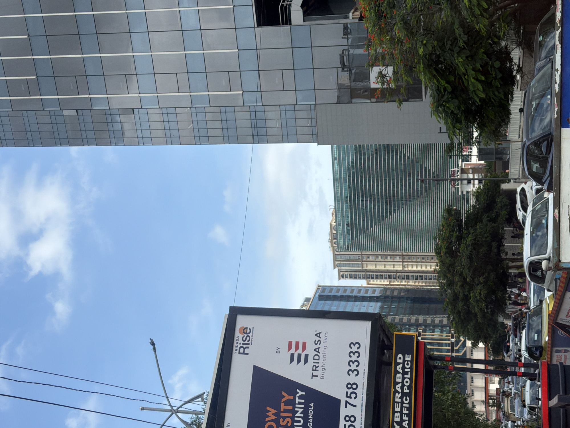 This is a vertical, eye-level, outdoor, daytime, full shot of a busy street scene in Hyderabad, India. The image is dominated by the bright blue sky and fluffy white clouds. On the left side of the frame, a billboard advertises "TRIDASA Rise" with the tagline "Where life shines." Below this, the text "CYBERABAD TRAFFIC POLICE" is visible on a yellow and black sign. A multitude of cars are visible, suggesting a traffic jam. In the background, several modern, glass-fronted skyscrapers stand tall, reflecting the sky. A large tree with red flowers is present in the lower right corner, adding a splash of color to the scene. The overall impression is one of urban activity and modern architecture.