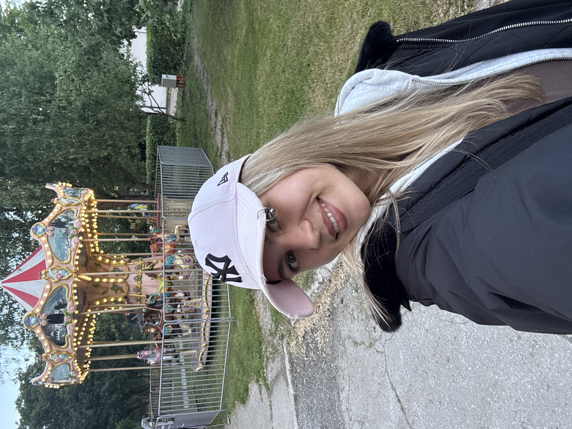 A young woman with blonde hair, wearing a black jacket, white hoodie, and a light pink baseball cap with a black "NY" logo, is taking a selfie in front of a carousel. The carousel is brightly lit with many lights and decorated with horses and other figures. It is surrounded by a metal fence. In the background, there are trees and grass. The location appears to be a park or amusement area in the city of Unknown, Unknown. A concrete path is visible on the ground near the woman.