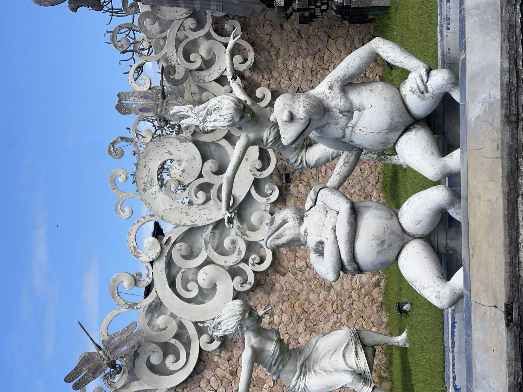 The image shows a vertical outdoor shot of statues located in Sukawati, Indonesia. Two humanoid figures with frog heads are stacked one above the other, their arms wrapped around their torsos. Adjacent to these figures are two female statues in flowing dresses, posed with outstretched arms and flowing hair. These statues are set against a background of rough, light brown stone tiles and are framed by ornate, swirling, metal scrollwork decorated with stylized dragonflies. The background transitions to include blue sky and grass. The statues are faded and appear to be made of concrete. A stone ledge with a weathered blue and white border supports the statues on the right.