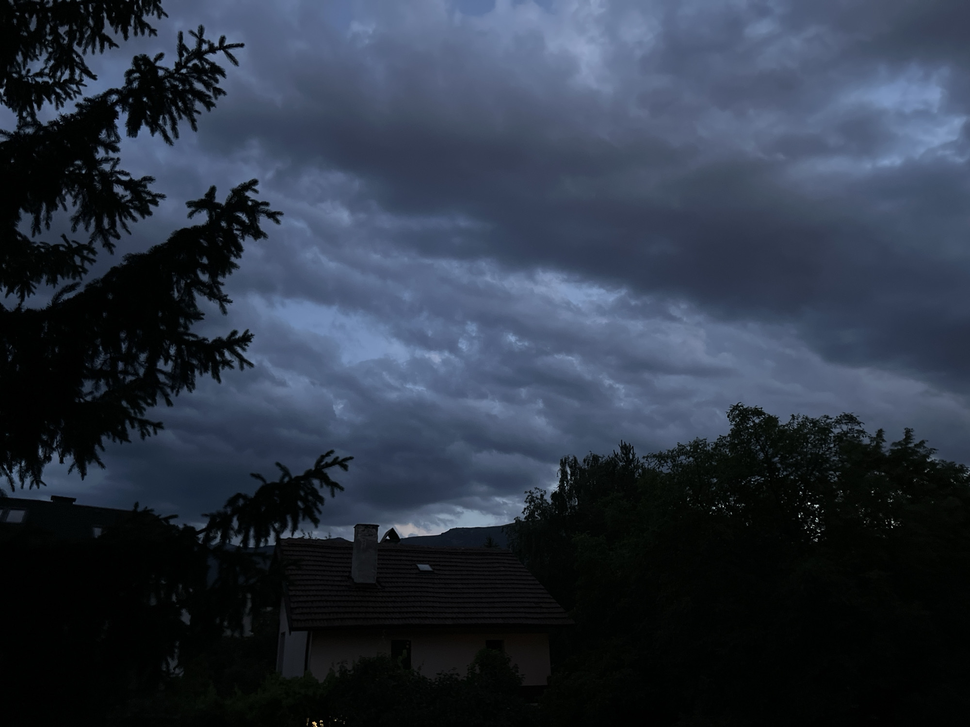 The image depicts a dark, cloudy sky over a residential area, likely in Sofia, Bulgaria, given the context. In the foreground, on the left, the dark silhouette of a fir tree branch stretches across the frame. Below and slightly to the right, a house with a reddish-brown tiled roof is visible. It has a chimney and a small window on the roof. The house appears to be partially obscured by dark trees and foliage. On the right side of the image, more dark trees form a dense line. In the distance, a dark, mountainous silhouette is faintly visible against the stormy sky. The overall atmosphere is somber and indicative of impending rain or twilight. No people or specific actions are depicted.