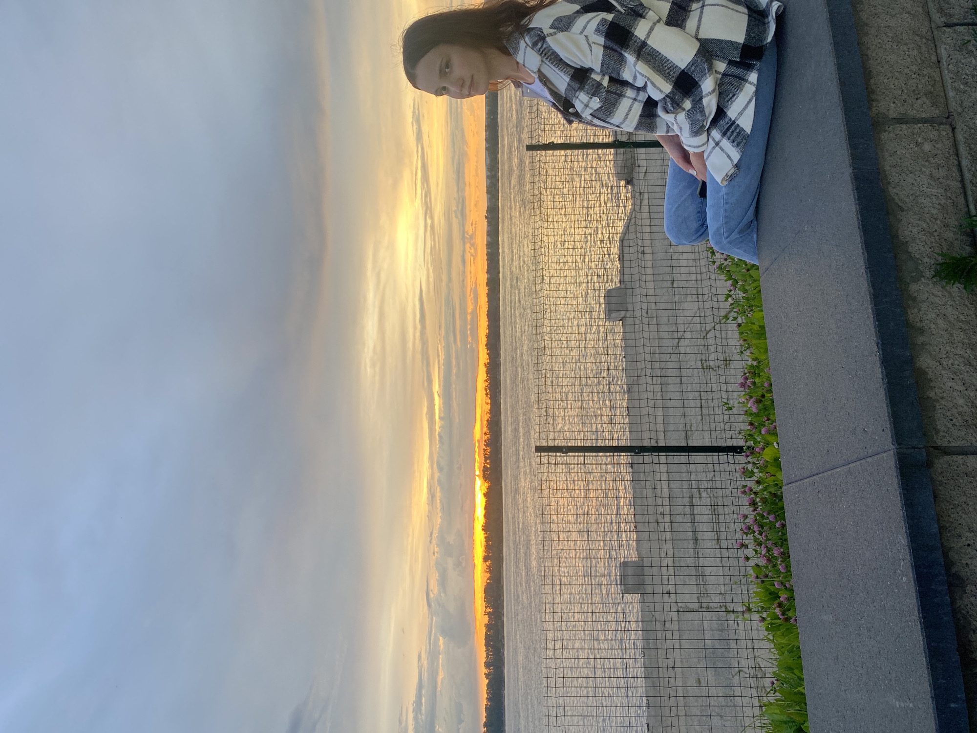 An outdoor, medium shot captures a young Caucasian female sitting on a concrete ledge with a river and sunset in the background. The female is positioned in the lower right quadrant of the frame, facing the viewer with a neutral expression. She has long, dark brown hair, and she is wearing a white t-shirt with a black and white plaid overshirt and blue jeans. The river is calm and reflects the orange and yellow hues of the sunset, which is low on the horizon. A line of trees is visible on the opposite bank of the river. A chain-link fence runs horizontally across the middle ground of the image, separating the concrete ledge from a sandy embankment. There are small pink flowers and green foliage growing along the edge of the ledge. The sky is filled with clouds that are illuminated by the setting sun. The scene appears to be a public waterfront area in Novokuybyshevsk, Russia.