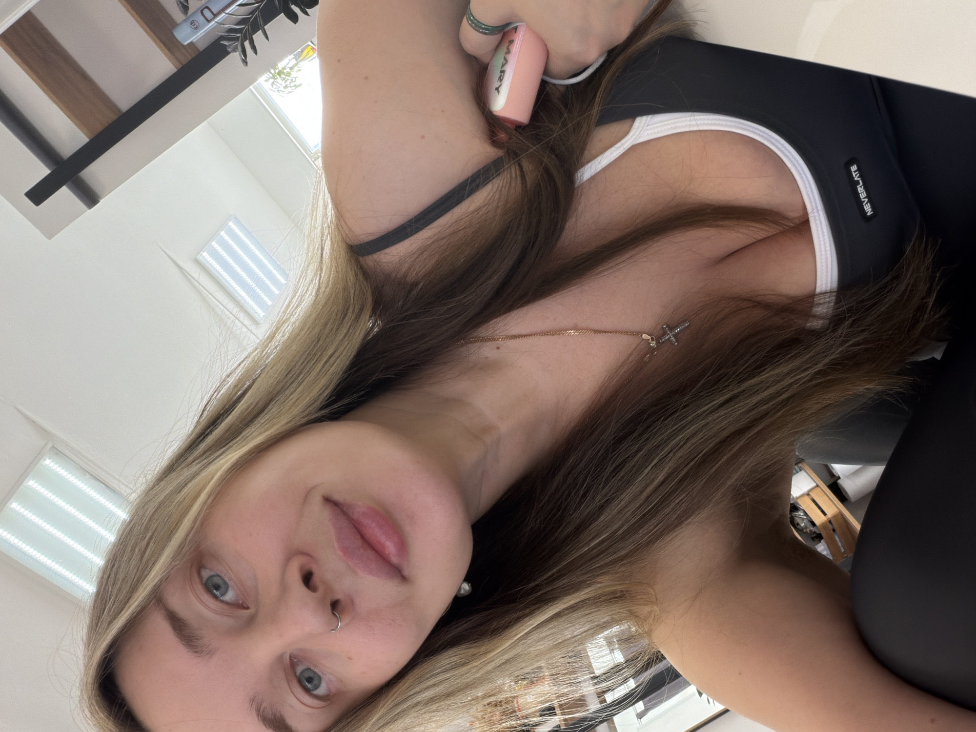 The image displays a rotated selfie of a young woman taken indoors, presumably in Chaikovsky, Russia. She is looking directly at the camera with a neutral to slightly relaxed expression. Her long, blonde and light brown hair frames her face, and she has blue eyes. A silver ring pierces her left nostril.
She is wearing a black top with white trim, possibly a sports bra or tank top, which features a small tag with the word "NEVERLATE" on it. A gold chain necklace with a small cross pendant is visible around her neck. On her left hand, she wears a greenish-blue ring, and a white bracelet or hair tie is on her left wrist. She holds a pink and white vape device in her left hand, with the word "MARY" visible on its side.
The background shows a brightly lit indoor space with white walls. Visible elements include a rectangular light fixture on the ceiling, a window with horizontal blinds, and parts of wooden furniture or shelving. A plant, possibly an olive branch, is also seen in the upper background. The bright lighting indicates it is daytime.
