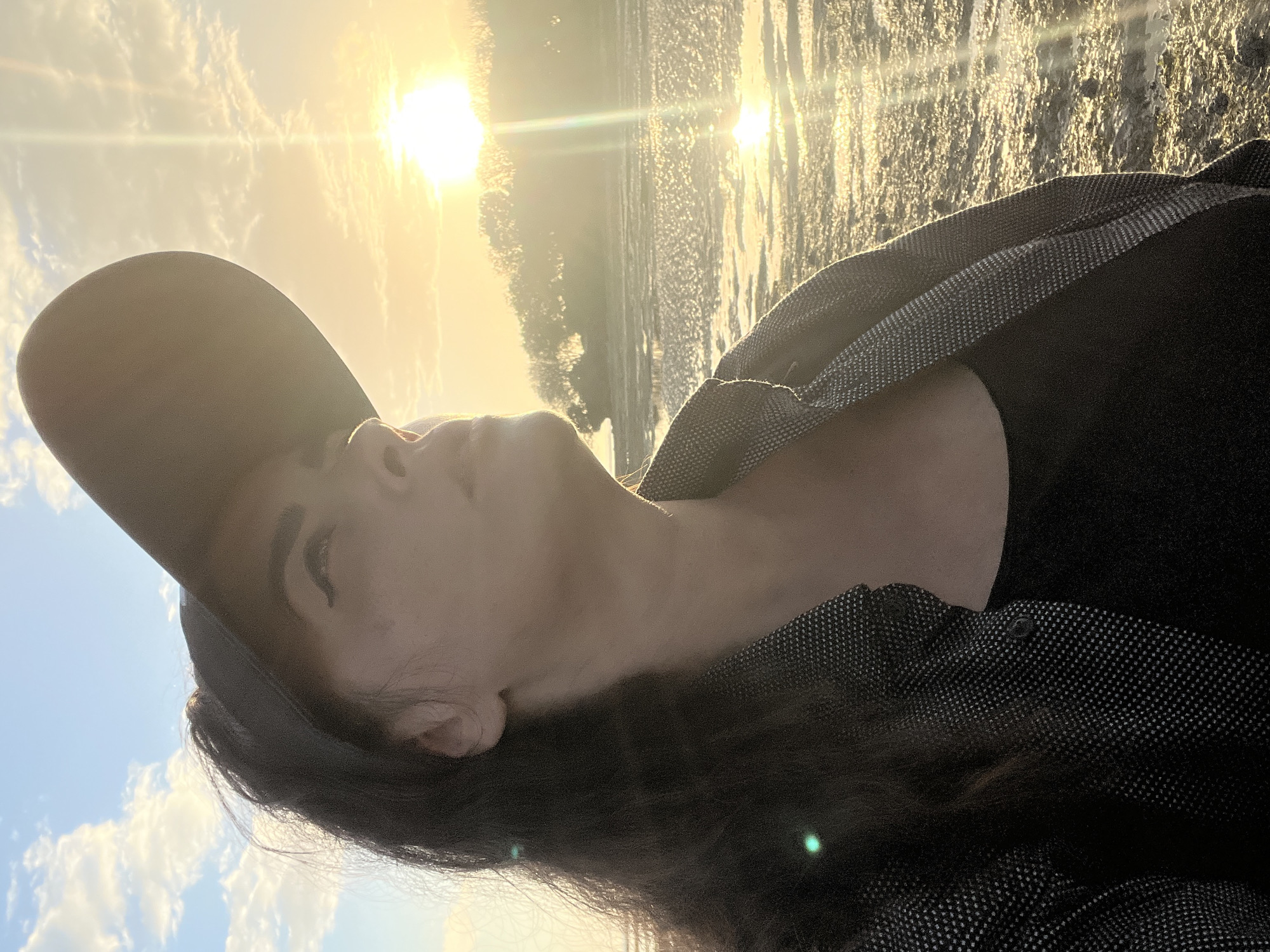 A person is captured in a close-up, low-angle shot at what appears to be a beach during sunset. The person is wearing a black baseball cap, a dark top, and a patterned jacket. They are looking upwards towards the sun, which is low on the horizon and casting a warm glow. The sun's rays are creating a lens flare effect in the image. The background shows a body of water, possibly the ocean, with a faint treeline in the distance. The ground in the foreground is sandy or muddy, with some patches of water reflecting the sunlight. The overall atmosphere is serene and golden, characteristic of a sunset. This scene could be Kuta Selatan, Indonesia, known for its beaches and vibrant sunsets.