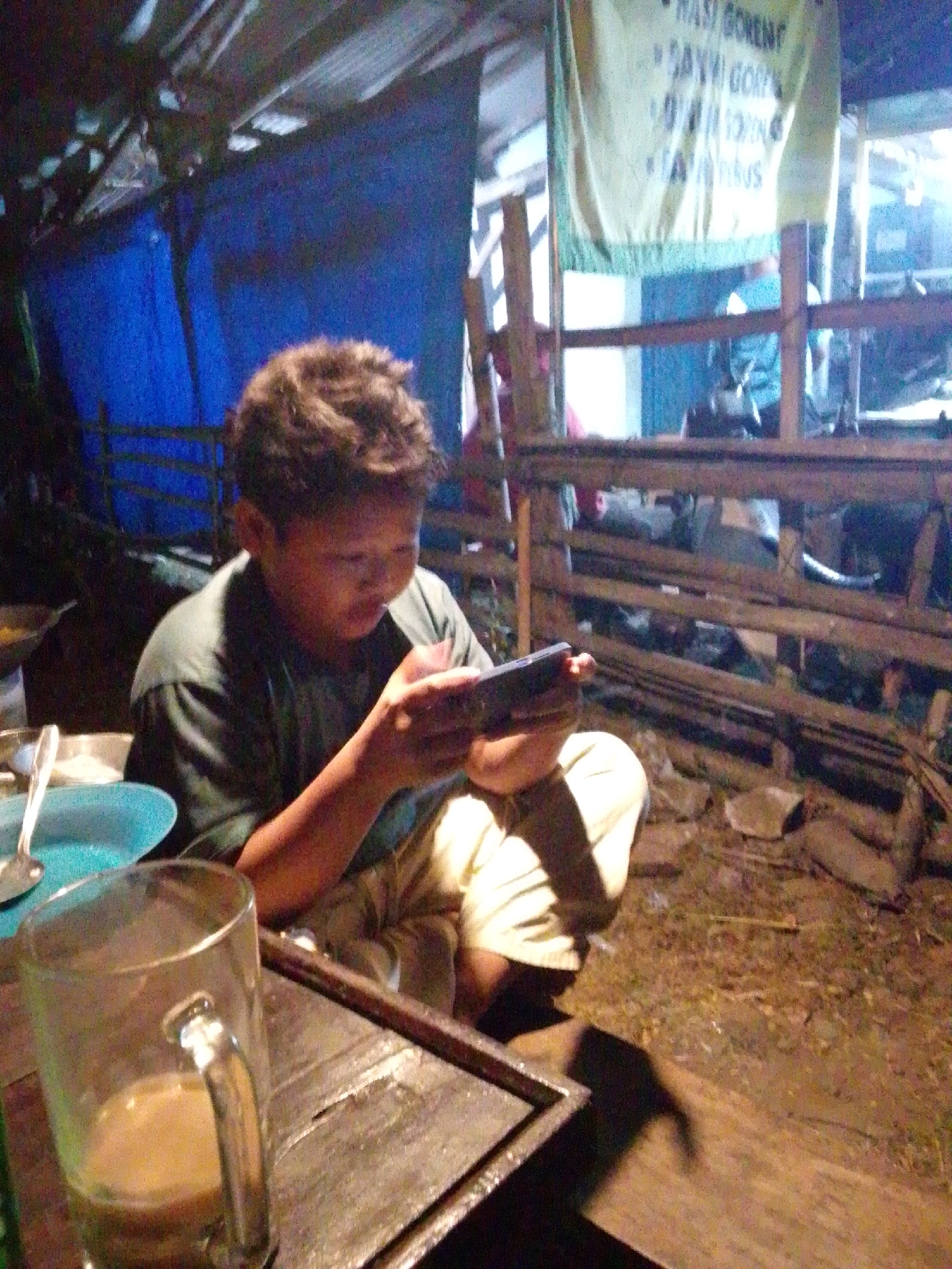 The image shows a young boy sitting on the ground, holding and looking at a smartphone. He is wearing a dark green t-shirt and light-colored pants. In the foreground, a clear glass mug with a light brown liquid is visible on a wooden table. To the left, a blue plate with a spoon is also on the table. The background appears to be an open-air structure with a corrugated metal roof and bamboo supports. A blue tarp is visible behind the boy. A banner with text, possibly a menu, hangs in the background. The ground is covered with dirt and straw. The overall scene suggests a casual, perhaps rustic, outdoor setting in Wirun, Indonesia.