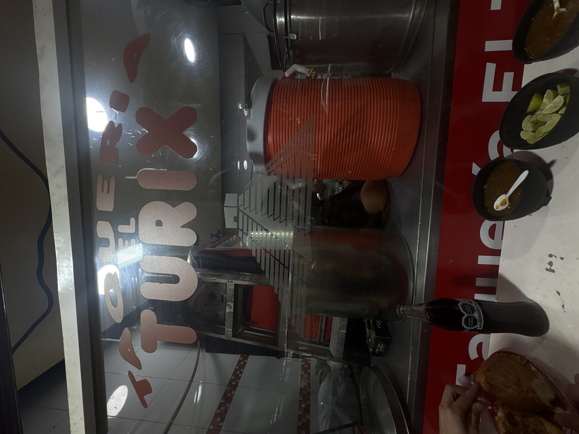 The image captures a street food vendor stall in Mexico City, Mexico. In the foreground, a glass bottle of dark soda with a label containing Mexican nutritional information stands upright. To its left, a person's hands are visible arranging food in a red plastic basket, possibly tacos or tortas. To the right, three black bowls are arranged: the leftmost contains brown salsa with a white spoon, the middle bowl holds sliced limes, and the rightmost bowl contains brown salsa with a white spoon. In the background, a large, ribbed orange beverage dispenser is prominent. Above this, a sign with pink letters is affixed to the glass window, reading "EL TURIX". Reflections on the glass show interior lighting and tiled walls. The overall impression is that of a busy, casual eatery.