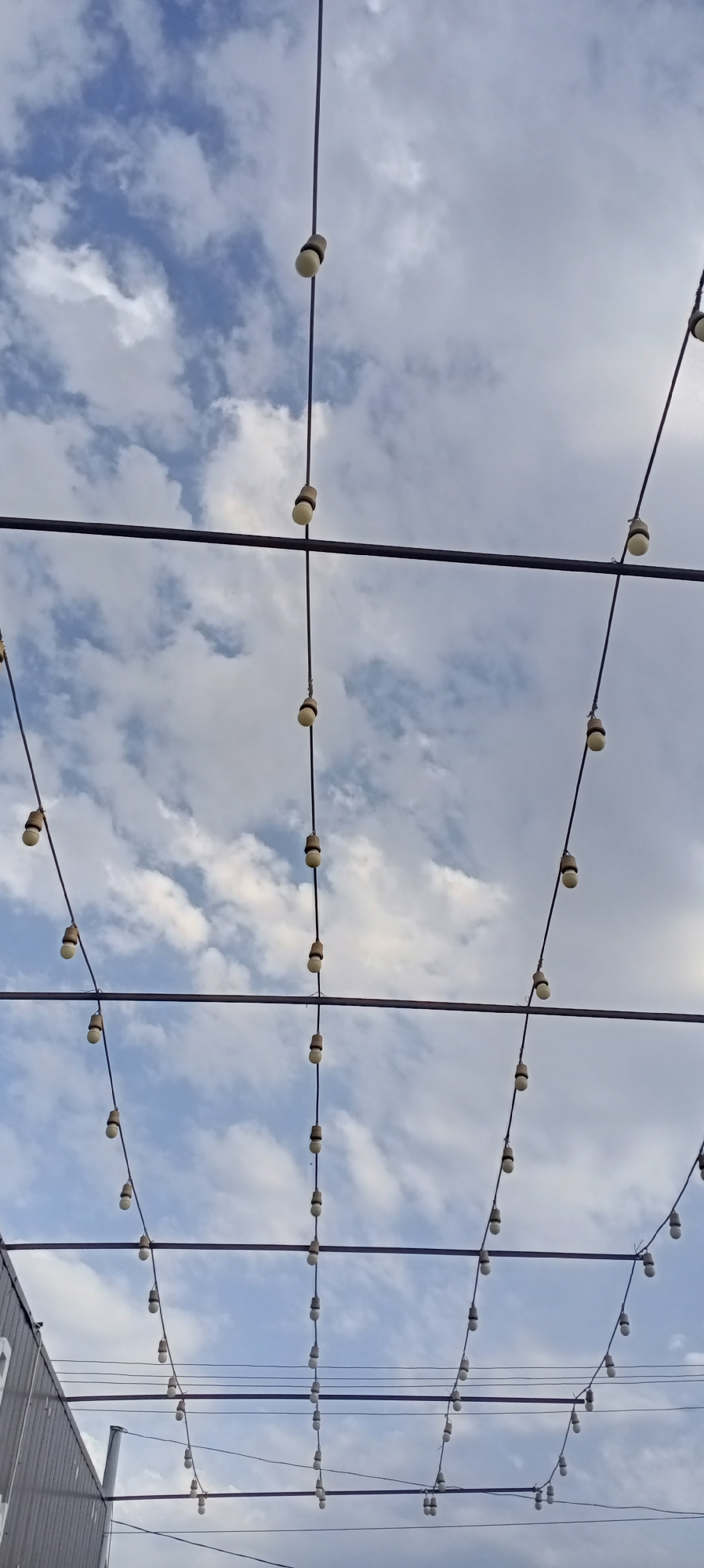 The image shows an outdoor scene with string lights suspended from wires against a cloudy sky. The lights are a mix of bulb and filament styles and are strung in a grid-like pattern. Some wires are attached to horizontal bars, creating a layered effect. In the lower left corner, a portion of a building with corrugated siding is visible. The overall setting suggests an outdoor event space or a decorated area in a city. The image does not contain any people or specific landmarks to identify its precise location within Sokuluk, Kyrgyzstan.