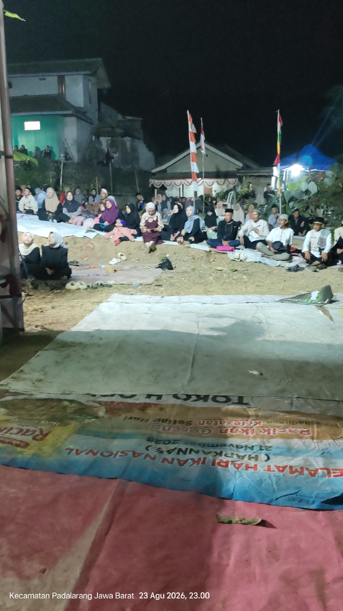 A crowd of people, predominantly women wearing hijabs and men, are gathered outdoors at night, sitting on the ground. They appear to be watching an event, possibly a religious or community gathering. In the background, there are simple buildings and tents, and national flags are visible. The ground is covered with tarps, and in the foreground, a banner with Indonesian text is partially visible, indicating "Kecamatan Padalarang Jawa Barat 23 Agu 2026, 23.00", suggesting the location and date of the event.