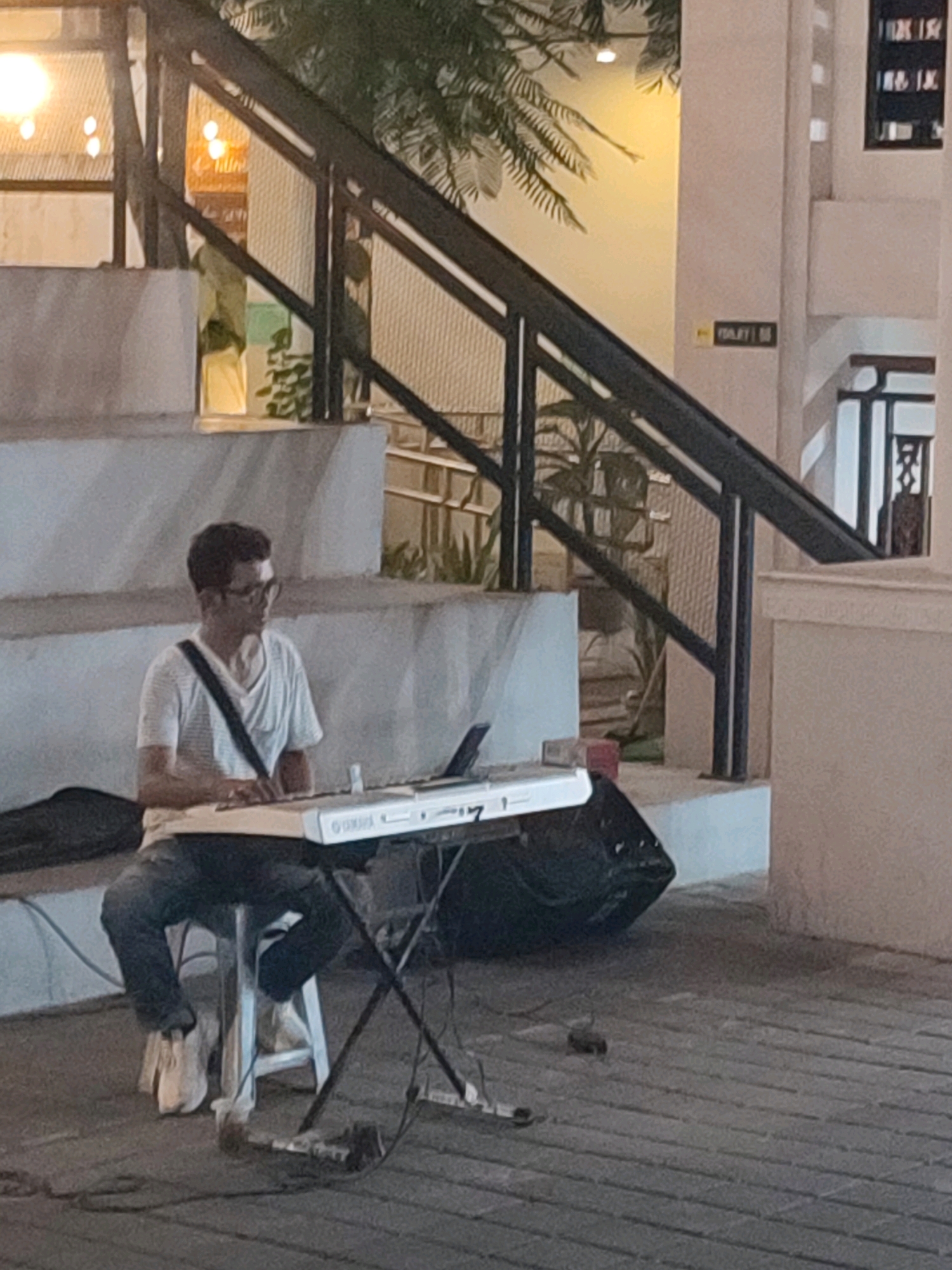A man with glasses, wearing a striped t-shirt and jeans, sits on a white stool playing a white Yamaha keyboard. He is positioned outdoors on a paved walkway, with a black speaker to his right. Behind him, a set of concrete stairs with a black metal railing leads up to a building. The lighting suggests it is dusk or evening. There is a small sign on the wall behind the railing that says "TOILET". The scene appears to be in a public area, possibly a plaza or entertainment district, in Yogyakarta, Indonesia.