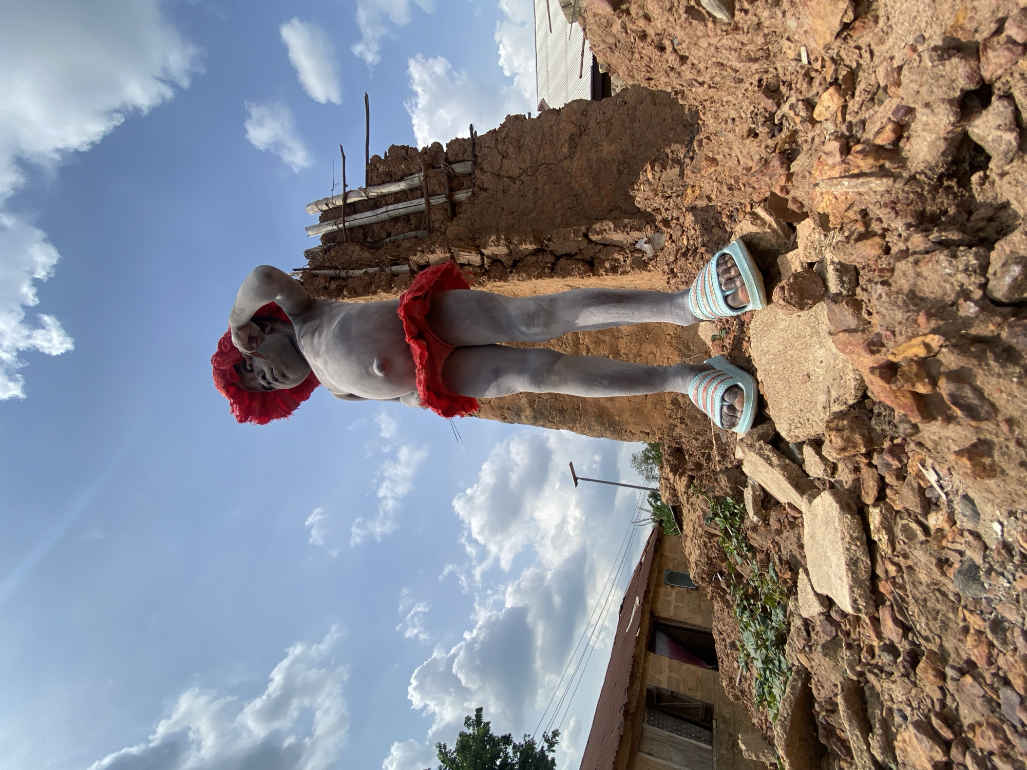 A grey-skinned individual wearing a red head wrap, a red garment around the waist, and blue sandals is seen standing on a rocky, uneven surface. The person appears to be near a weathered mud wall structure. In the background, there is a clear blue sky with scattered white clouds and a building with a rusted roof. The photograph is taken from a low angle and seems to be taken in a rural setting, Igbarra, Nigeria.