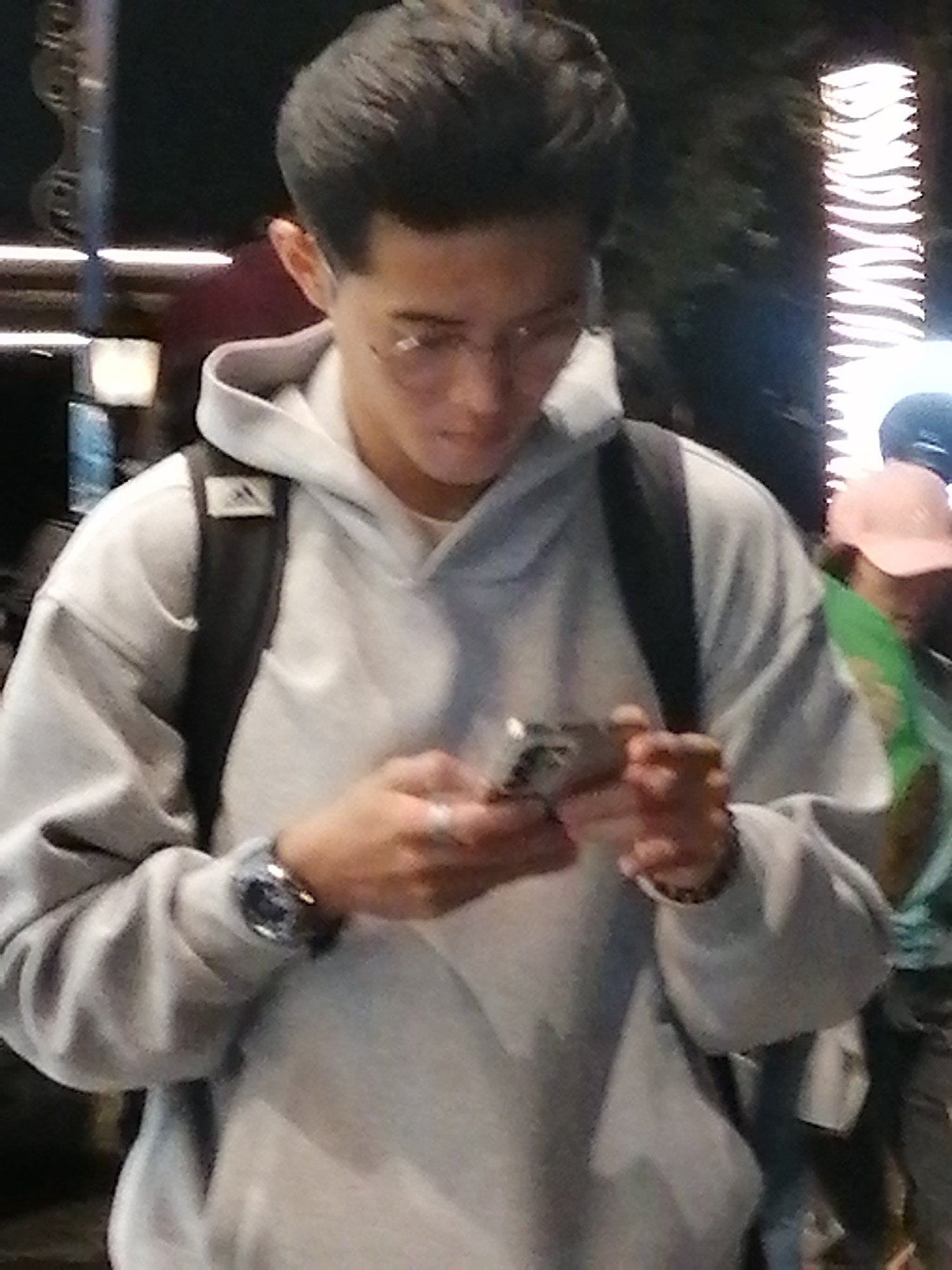 The image depicts a young man wearing a light gray hoodie and a black backpack, looking down at his mobile phone. He has short, dark hair and appears to be wearing glasses. On his left wrist, he has a watch with a dark band and a metallic face. The background is blurred and dimly lit, with some indistinct vertical lights suggesting an urban environment at night. To the right of the main subject, a person wearing a pink cap and a green shirt is partially visible. The scene does not provide specific visual cues to pinpoint its location within Da Lat, Vietnam, beyond suggesting an outdoor, possibly public, setting at night.